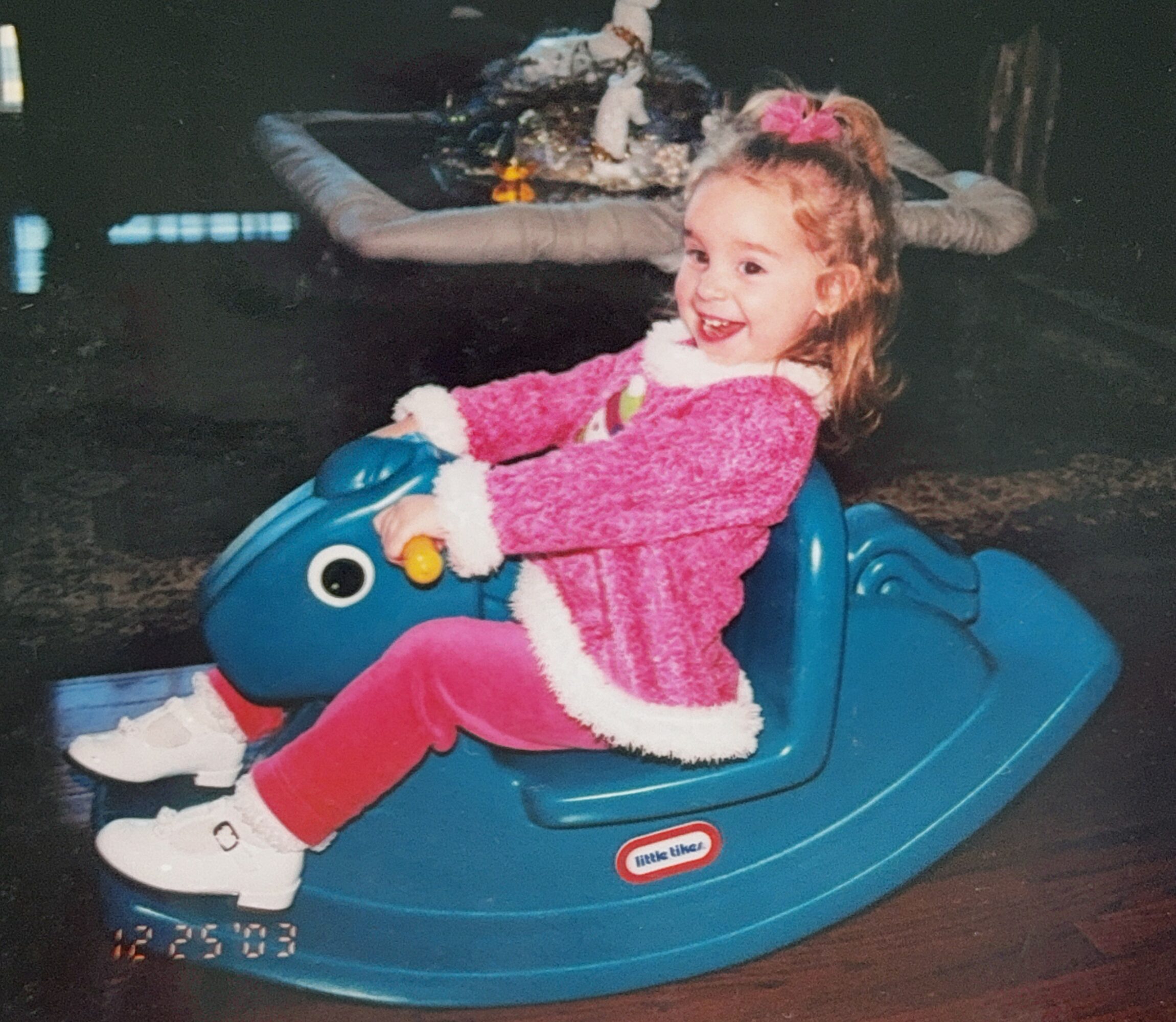 Young girl with curly hair in a pink sweater and pants rides a blue rocking toy with a face, smiling.