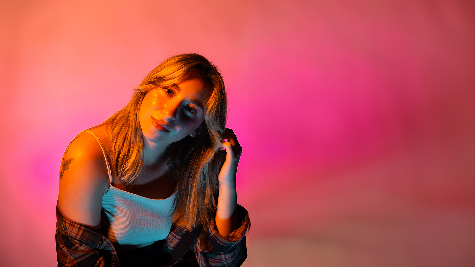 Young woman with blonde hair, wearing a white top, posing against a pink and purple background.