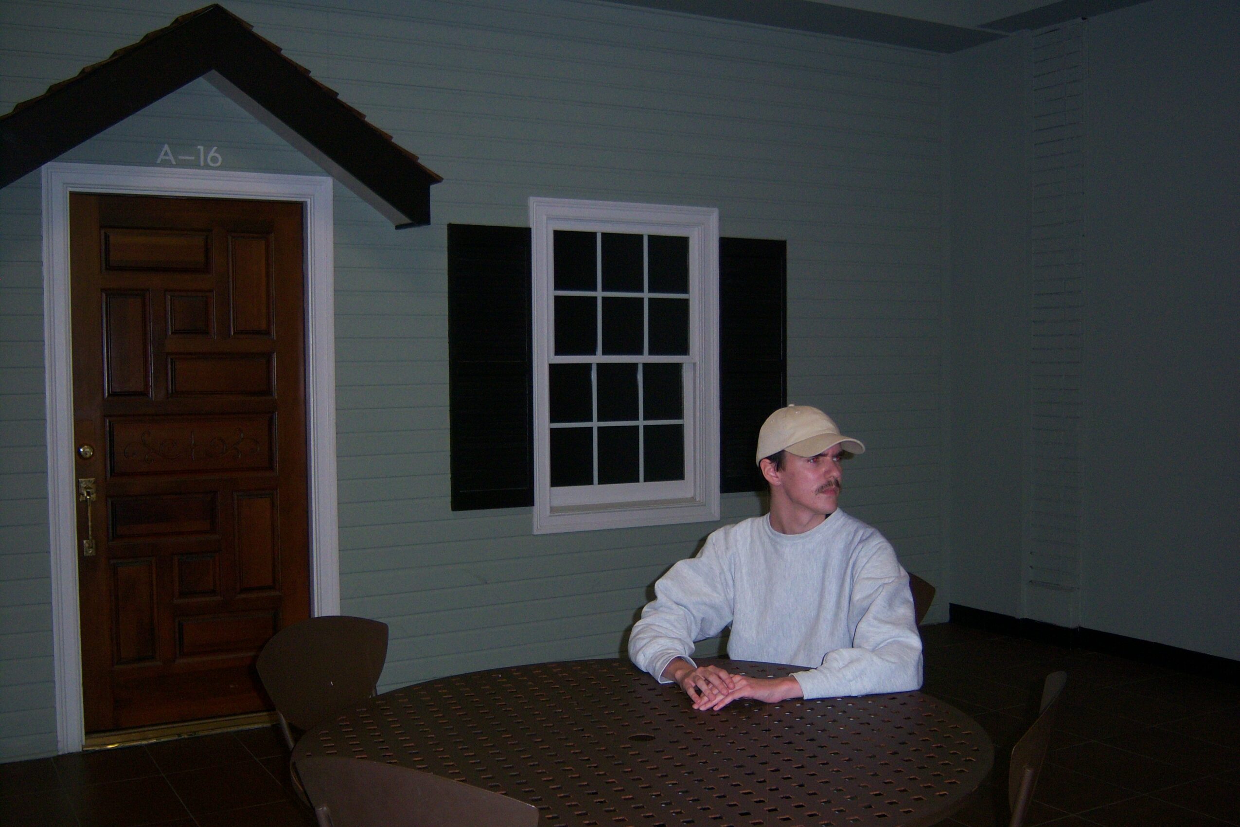 Person sitting at a round table in a room with a window and a door, wearing a cap and white clothing.