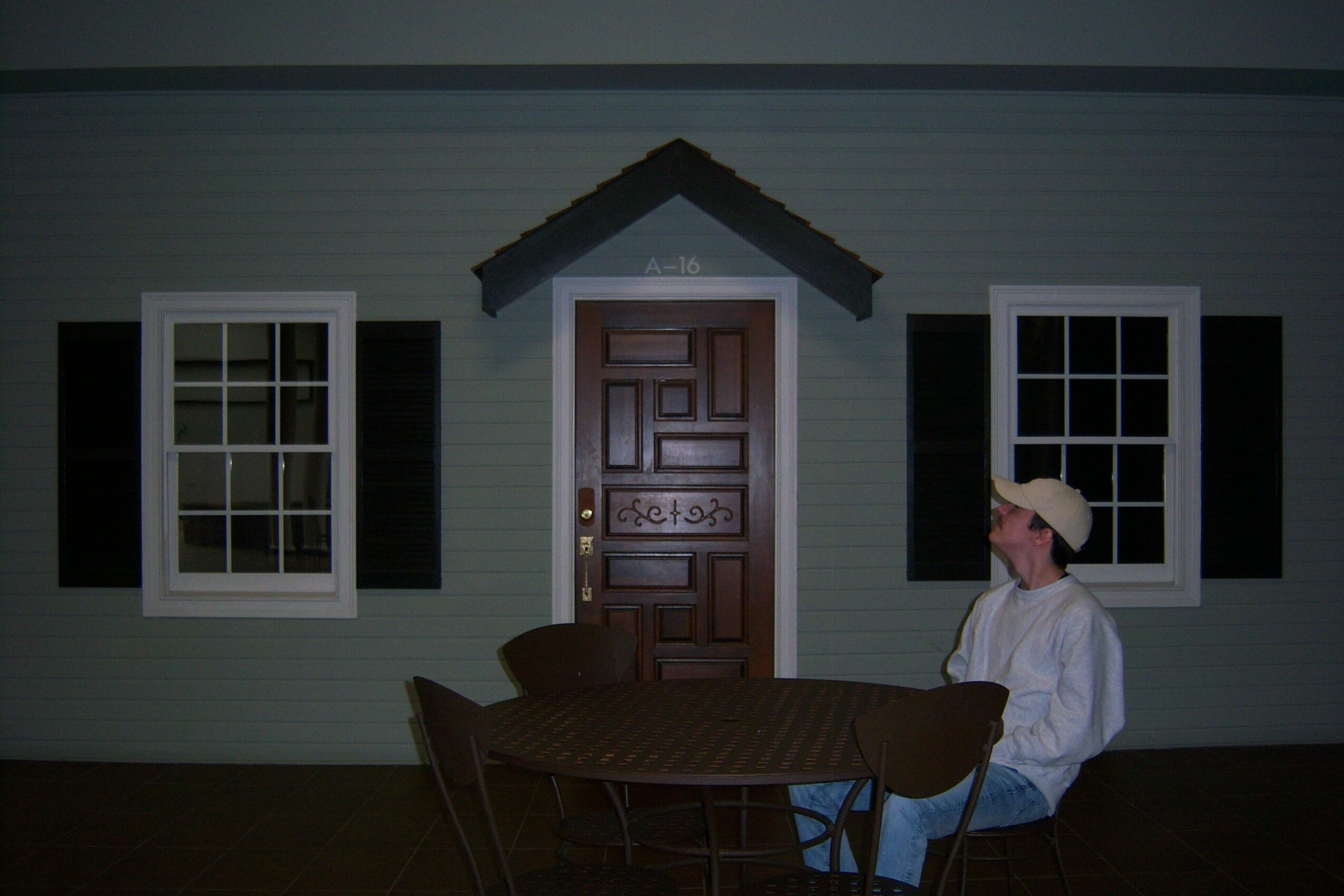 Person sitting at a table outside a house with two windows and a door, wearing a cap and white shirt.
