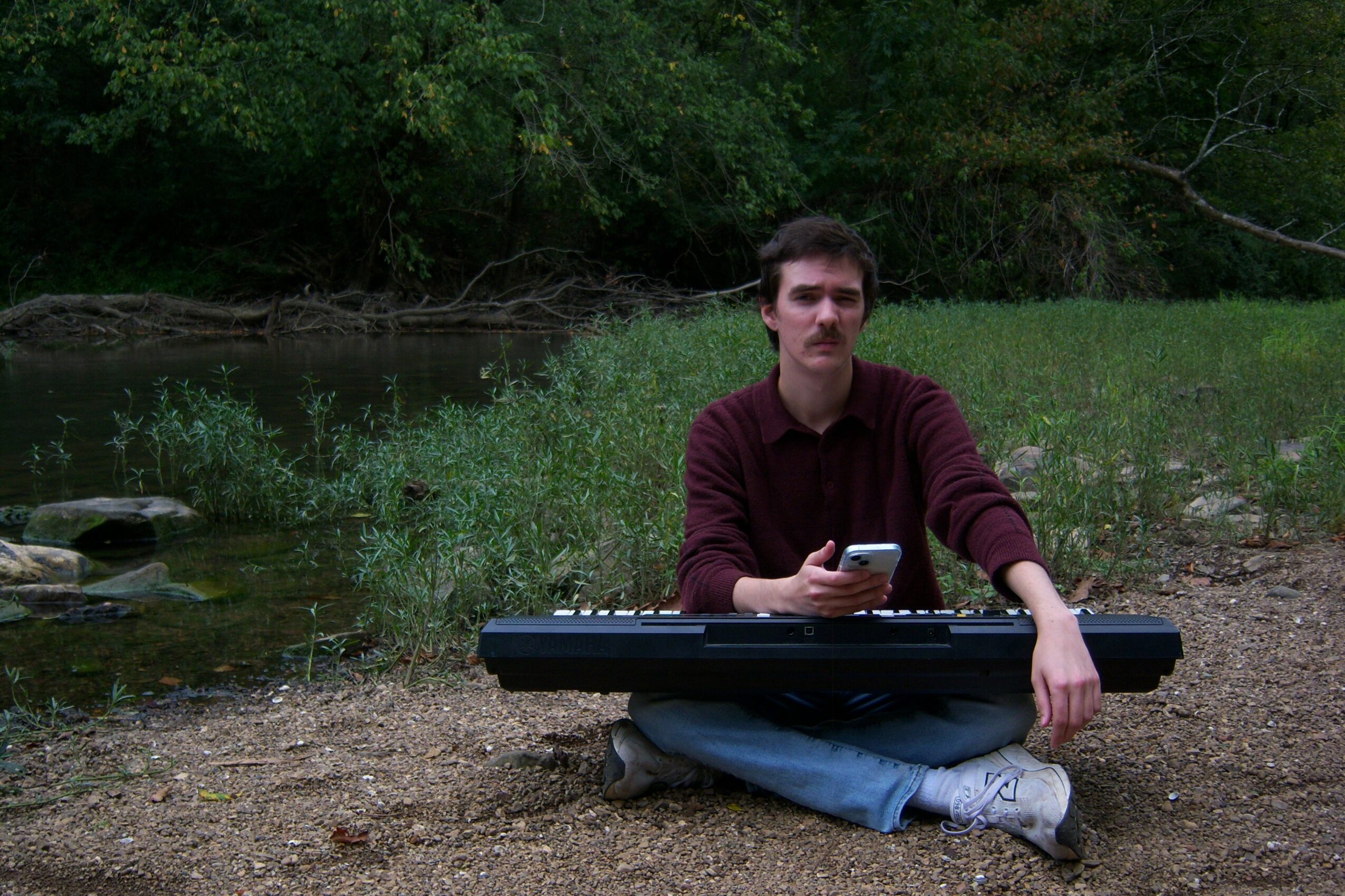 Person sitting cross-legged outdoors with a keyboard and phone, near a river and trees.