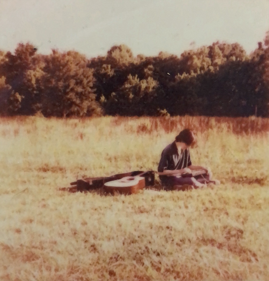 Person sitting on grass with a guitar and a book, in a field with trees in the background.