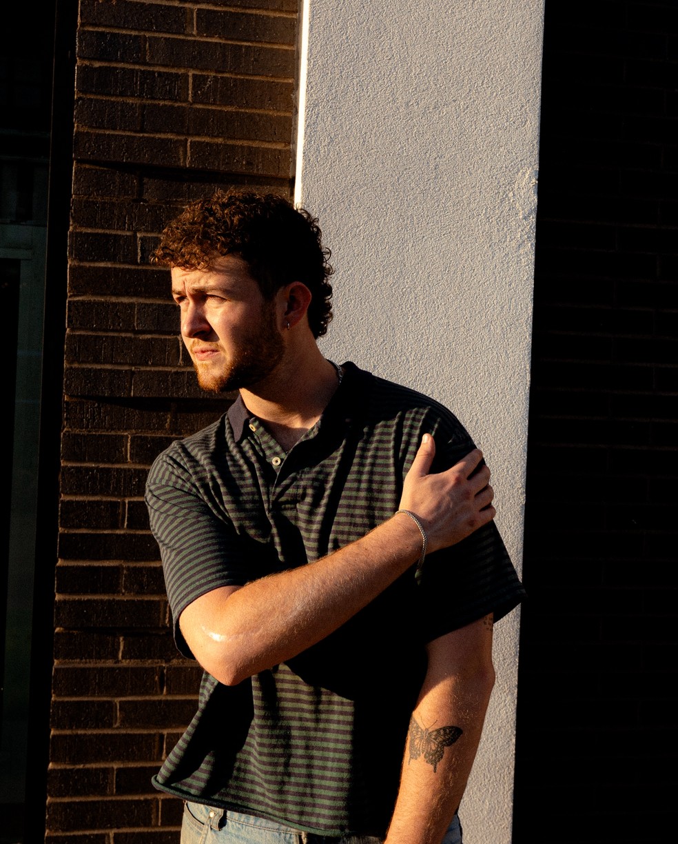 Young man with curly hair and beard, wearing a dark checkered shirt, standing outdoors in sunlight, leaning against a wall.