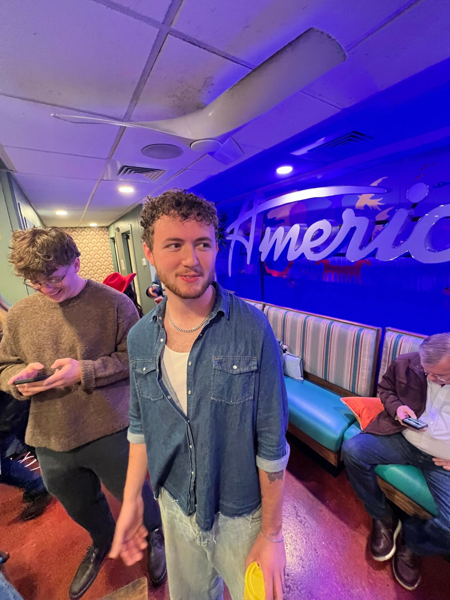 Young man with curly hair and denim jacket standing in a colorful indoor space with people and neon lights.