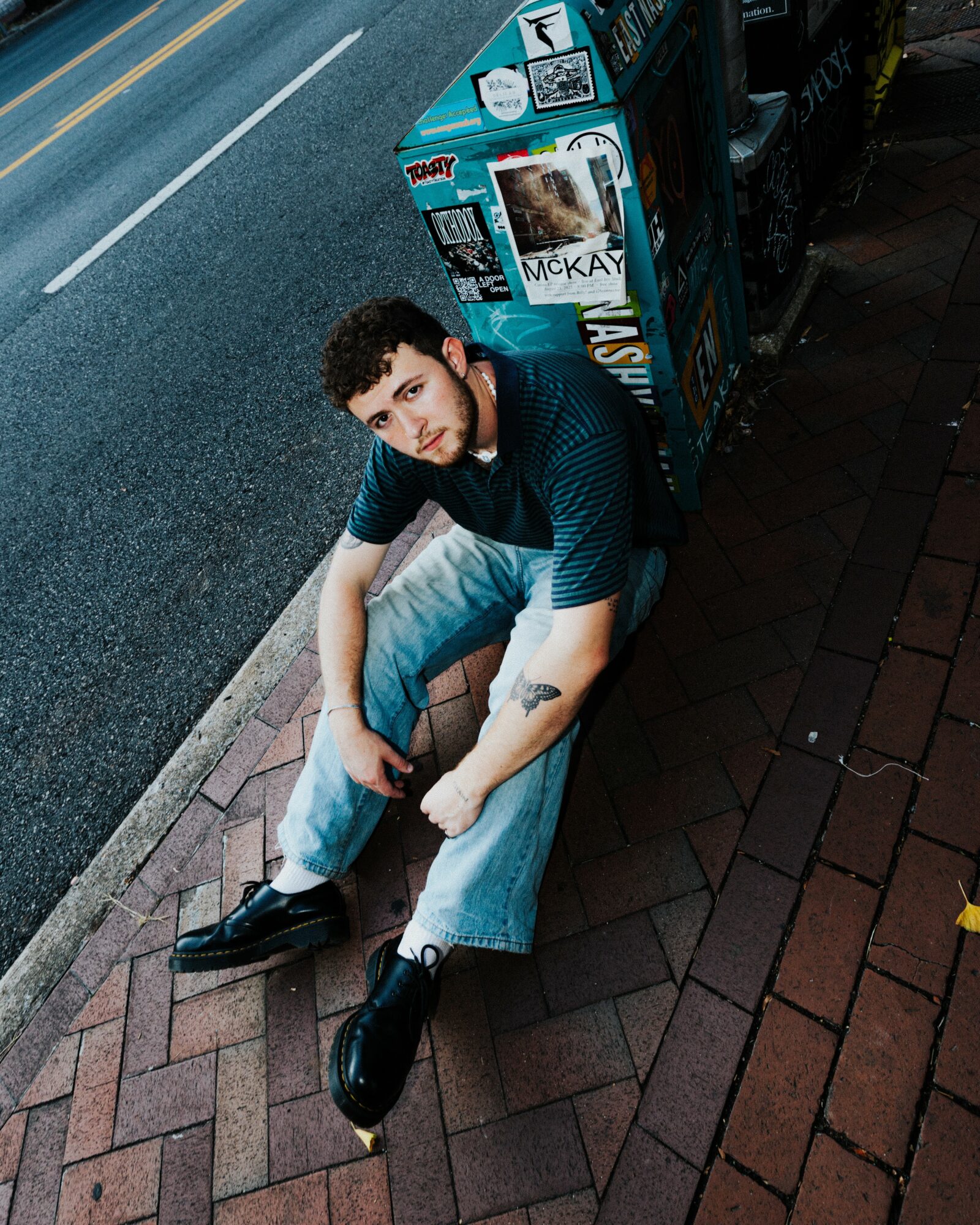 Young man with curly hair sitting on a brick sidewalk next to a street, looking at the camera, wearing a striped shirt and jeans.