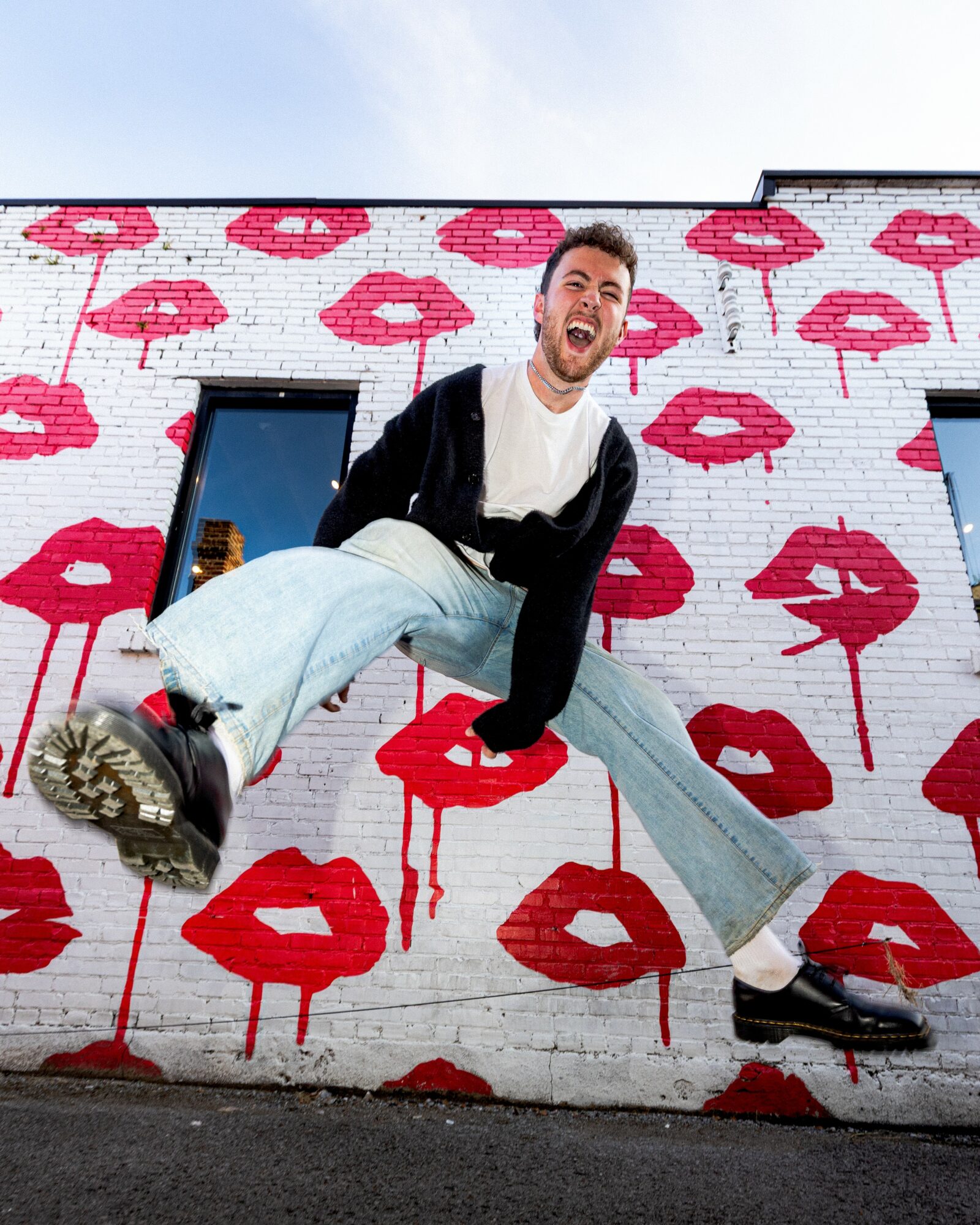 Person with a joyful expression leaning against a wall with pink lipstick kiss marks, wearing casual clothing and sneakers.