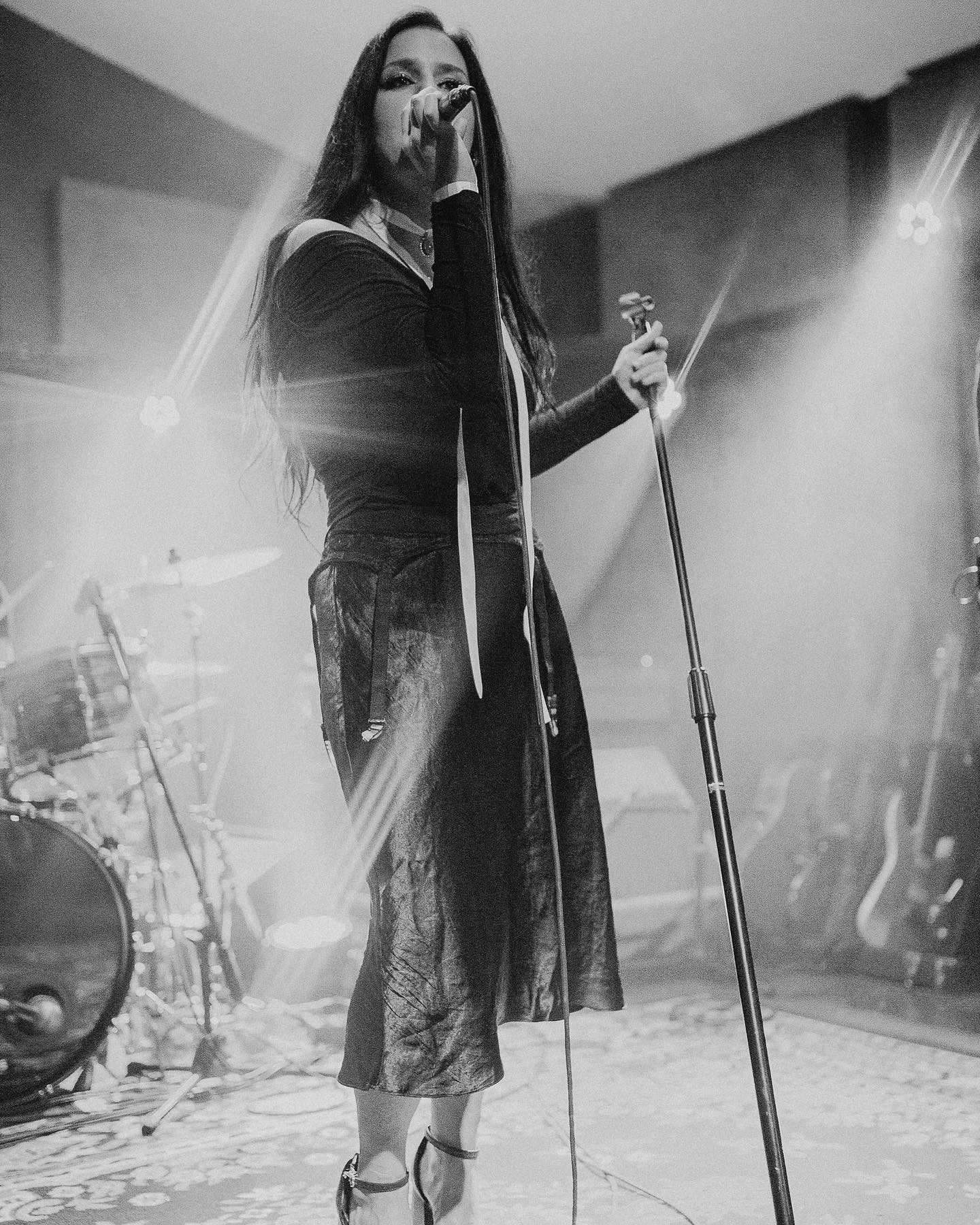 A woman singing into a microphone on stage with musical instruments in the background, illuminated by stage lights.
