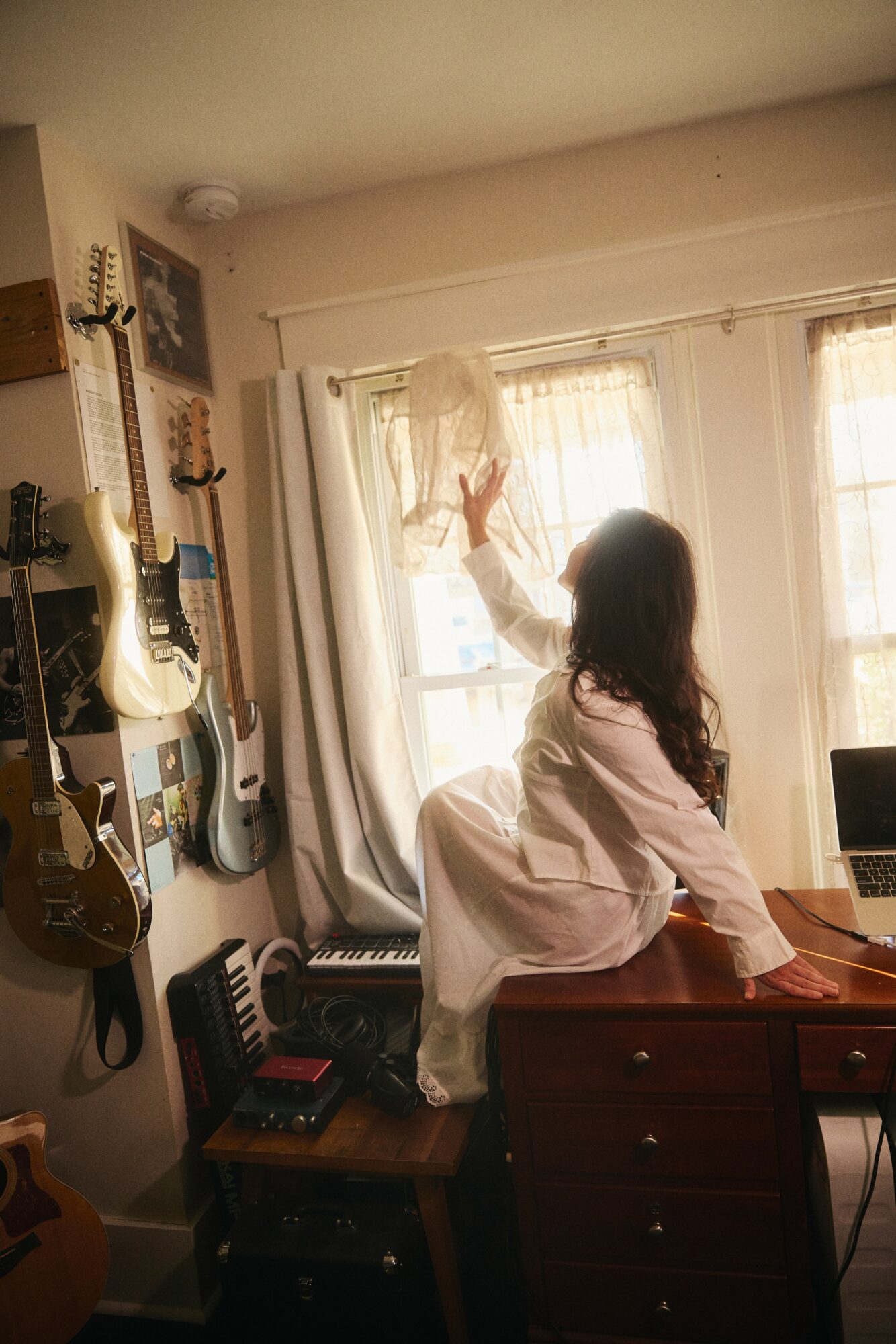 Person with long dark hair sitting on a dresser, reaching toward a window with sunlight, surrounded by musical instruments and equipment.