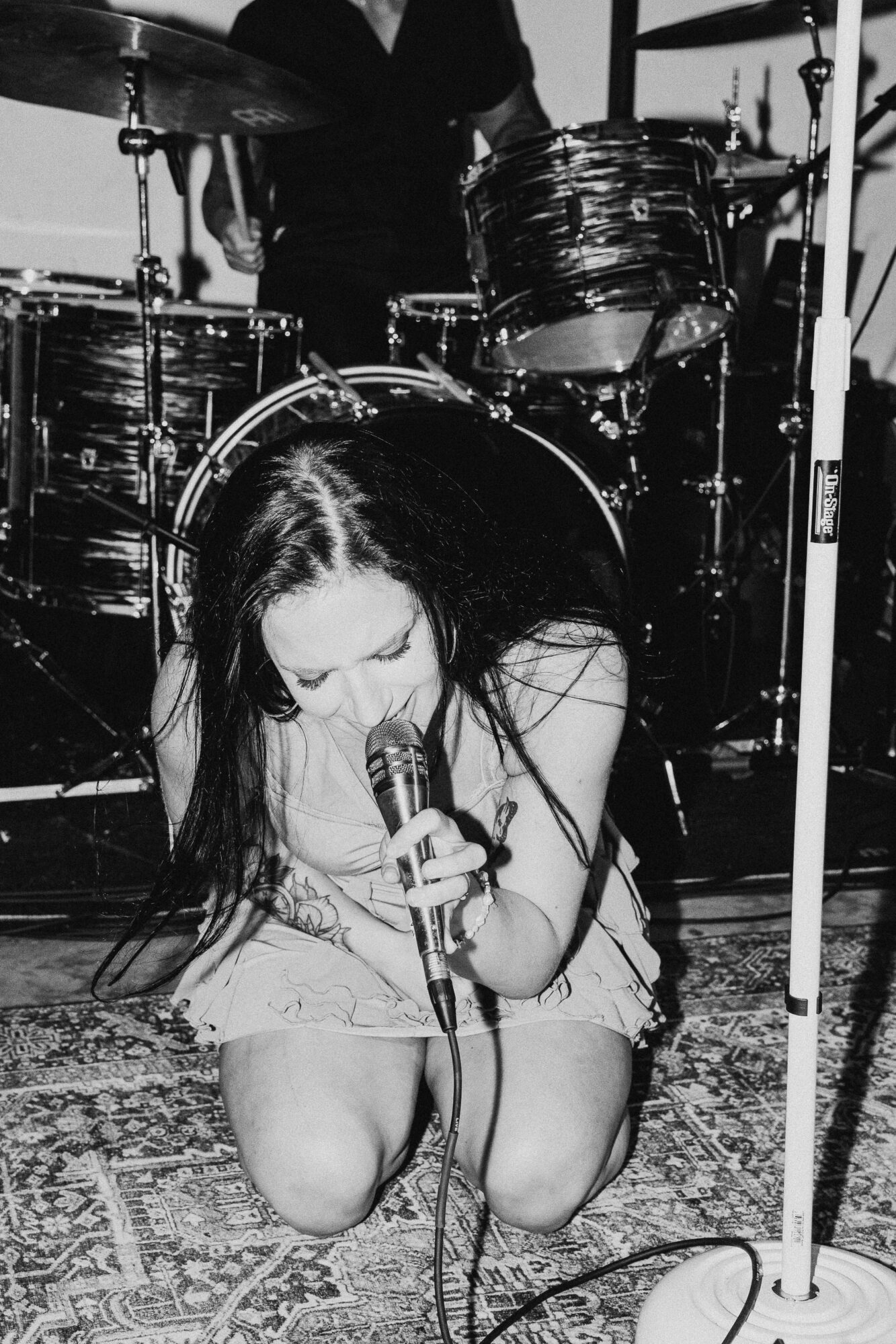 A woman with long dark hair kneels and sings into a microphone on stage, with drums in the background.