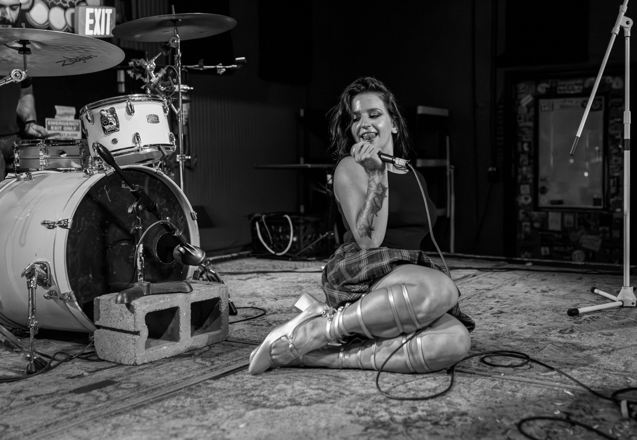 Woman sitting on stage with microphone, drums and equipment in background, smiling and singing, in a music venue, black and white.