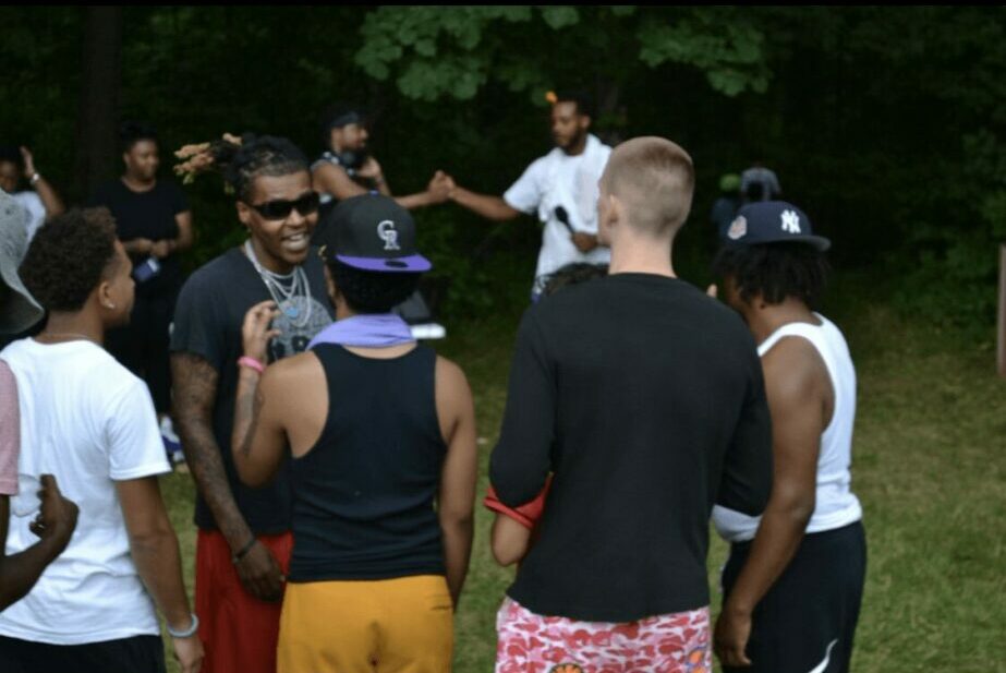 Group of people outdoors listening to a person speaking, surrounded by trees and greenery.