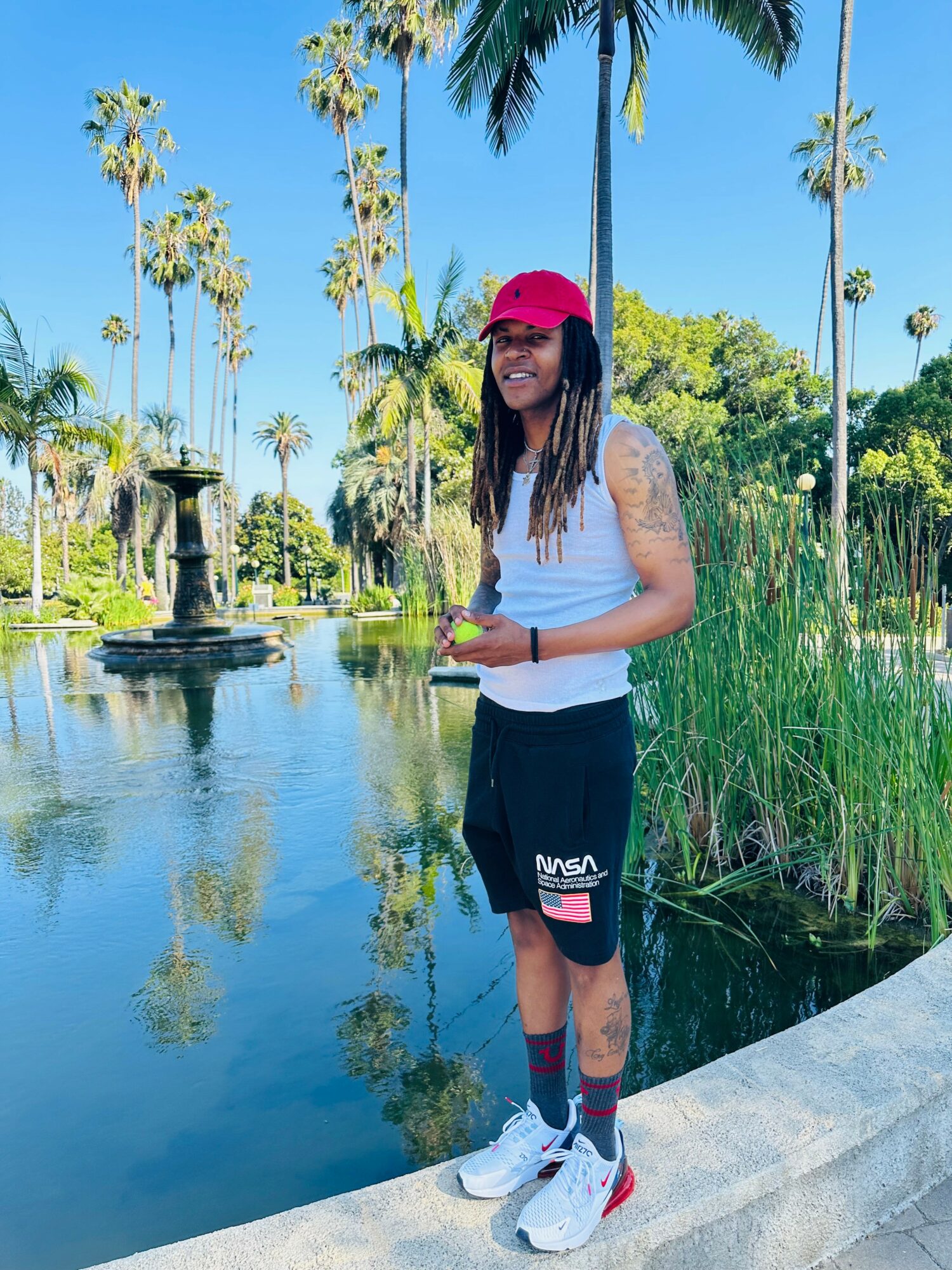 Person with long hair, wearing a red cap, white sleeveless top, black shorts, and white sneakers, standing by a pond with palm trees.