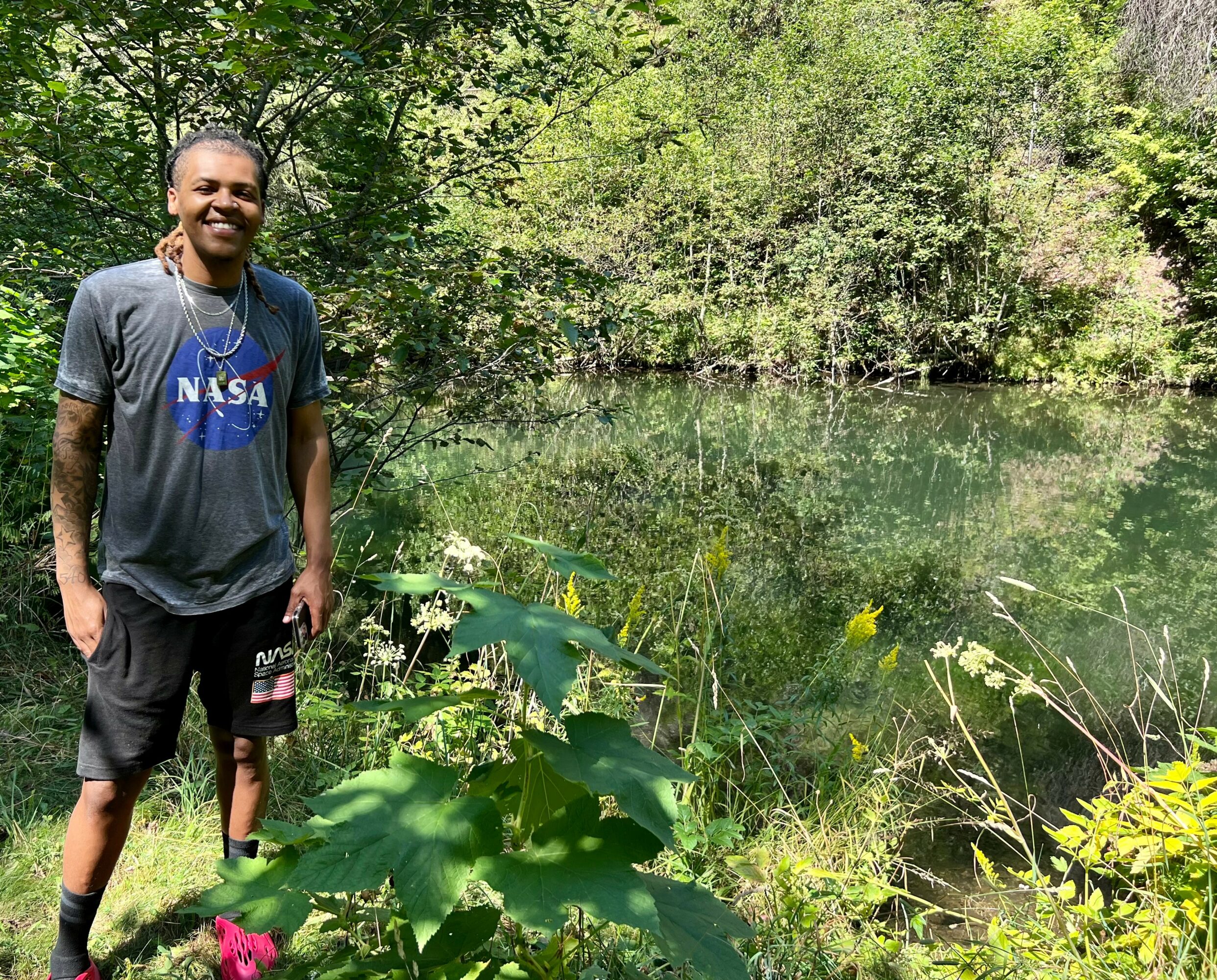 Smiling person standing outdoors near a body of water with trees and greenery in the background.