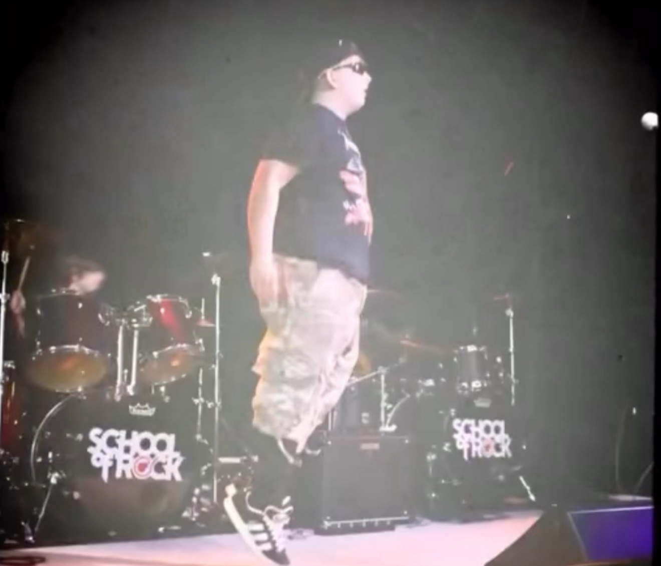Person standing on stage with drums and equipment, wearing sunglasses, a black T-shirt, and camouflage pants, performing or speaking.