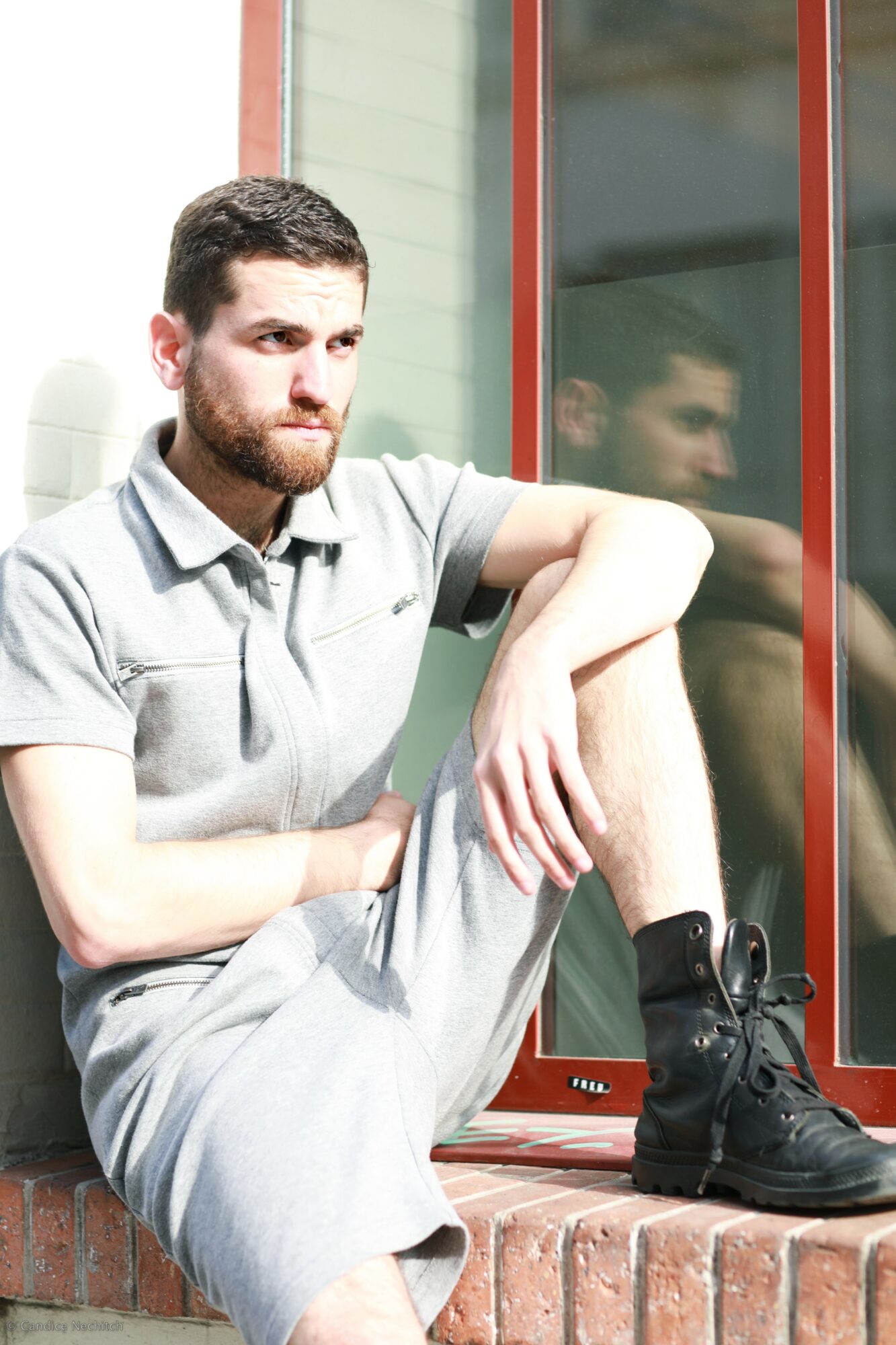 Young man with beard sitting outside, leaning against a wall with a window, wearing a gray polo shirt and shorts.