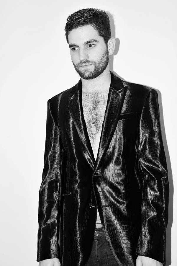 Man with dark hair and beard wearing a shiny black blazer, looking to the side, in black and white.
