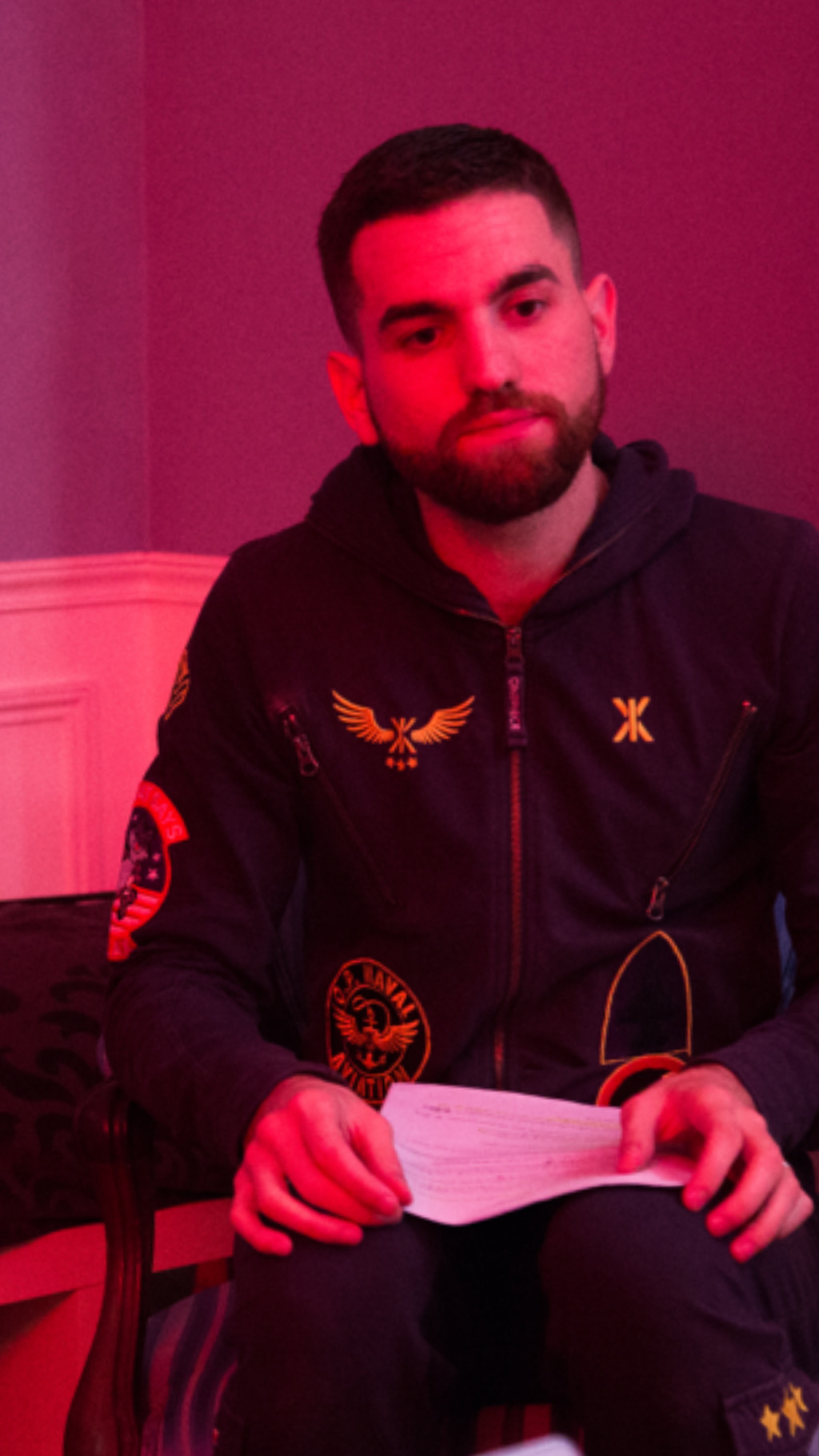 Young man with dark hair and beard, wearing a black jacket with patches, sitting indoors with pink lighting.
