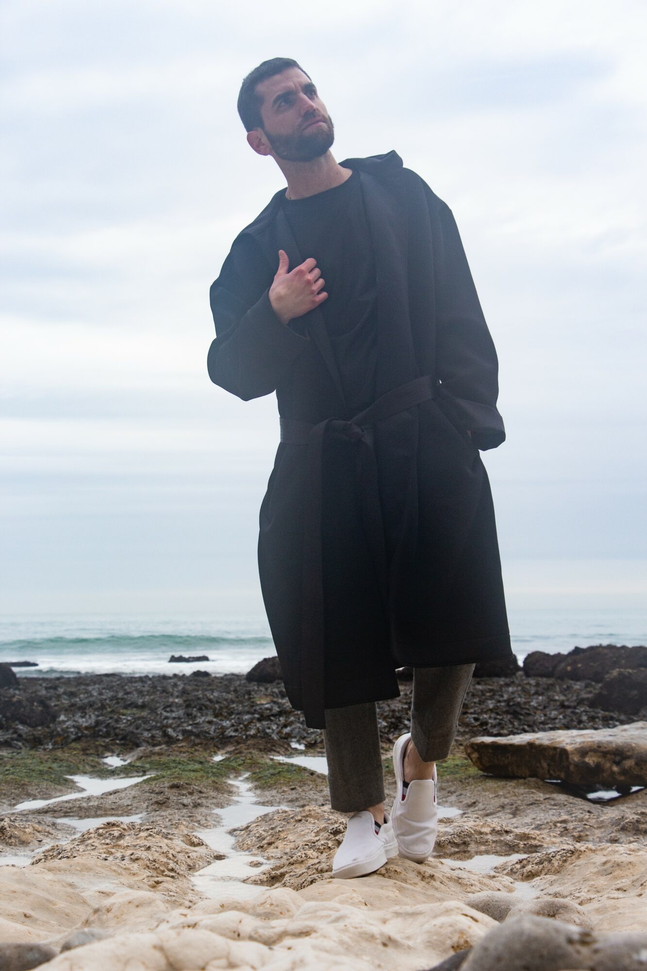 Person standing on rocky beach, wearing dark coat and pants, with hand on chest, against cloudy sky and ocean background.