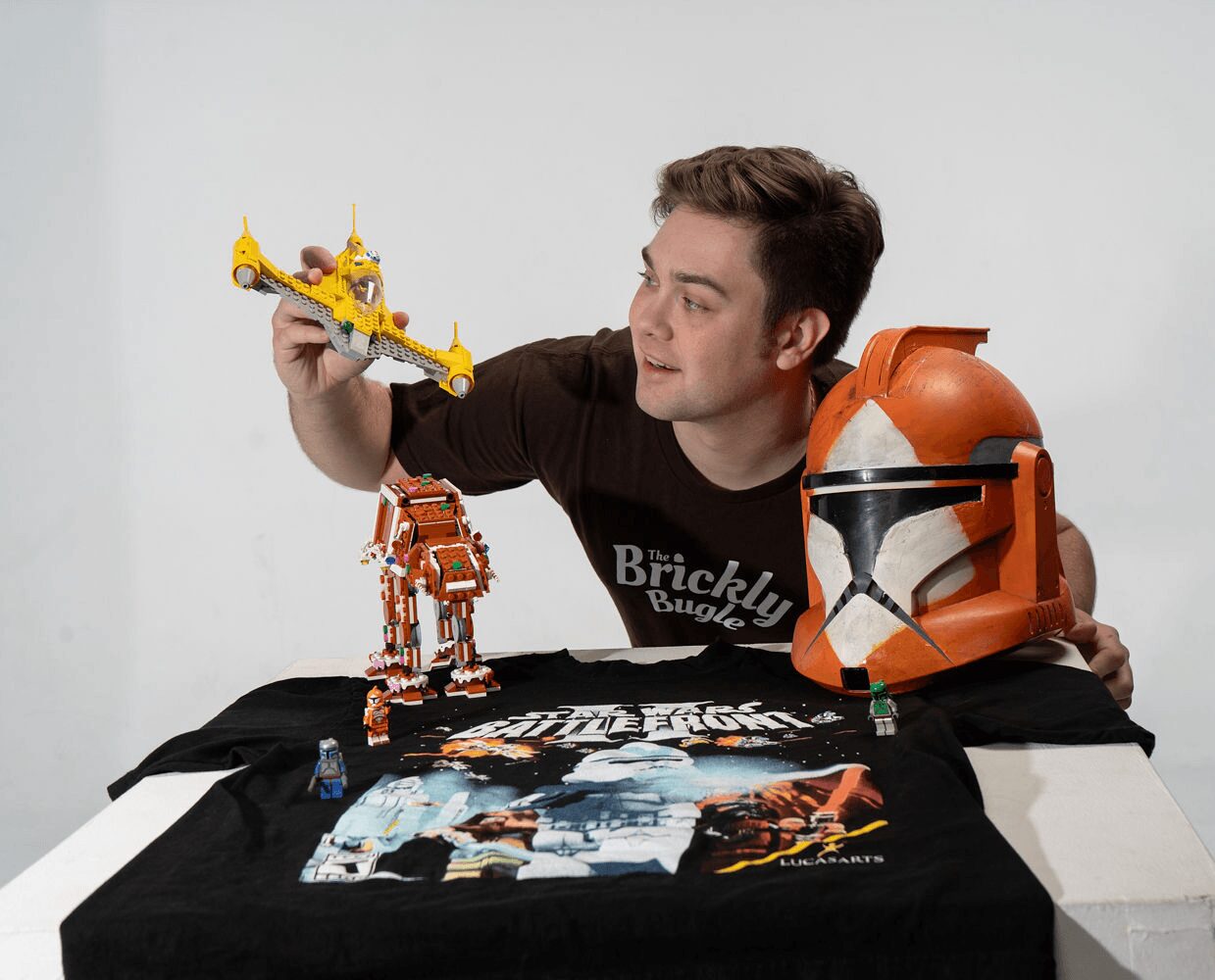 Man with a Bricklink T-shirt displays LEGO Star Wars figures and a poster on a table, holding a LEGO spaceship and a helmet.