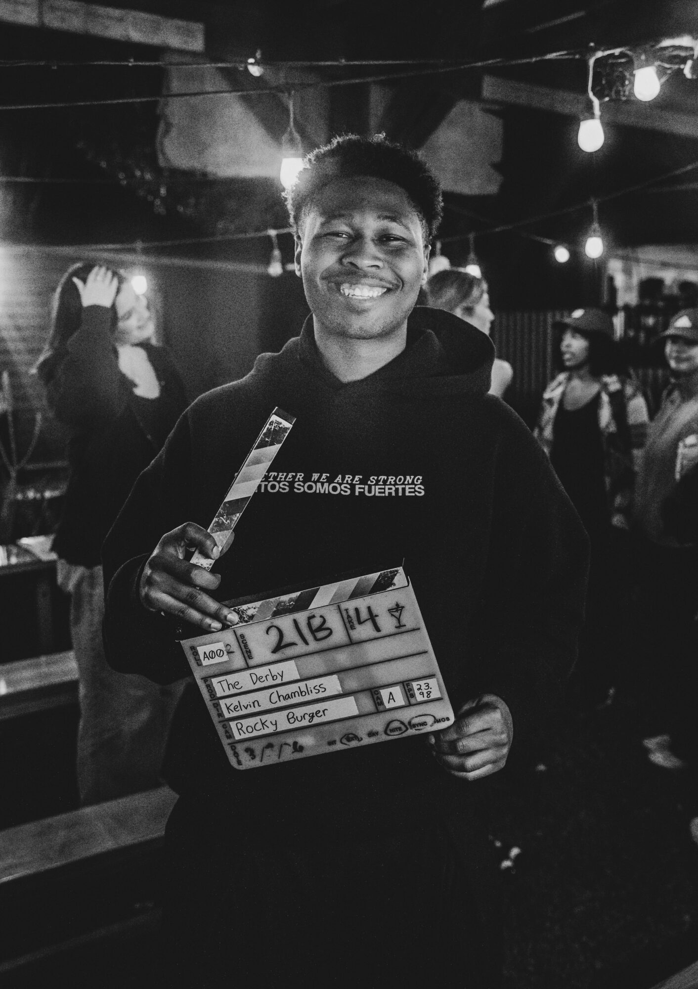Person smiling holding a clapperboard in a dimly lit room with other people in the background.