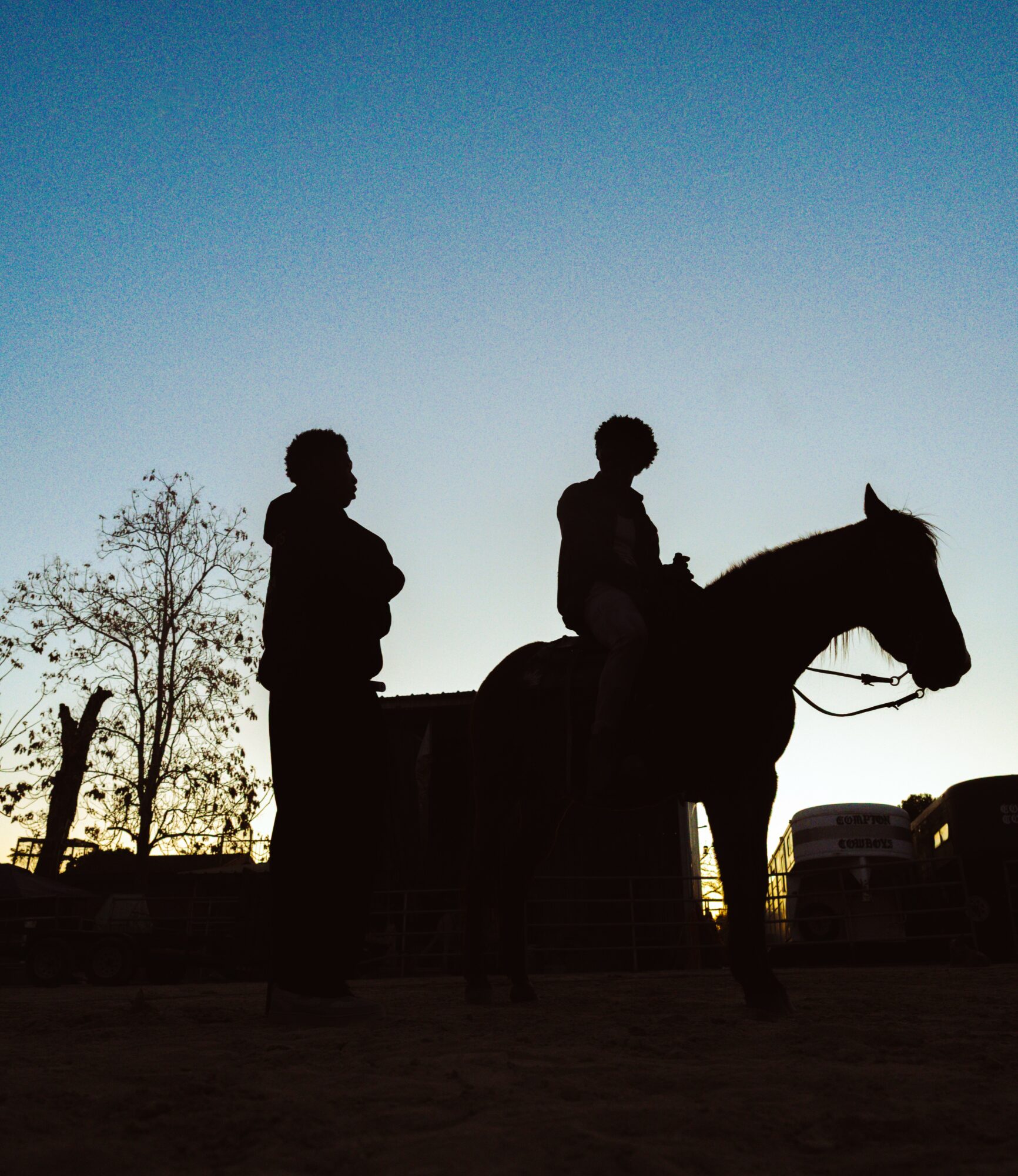 Silhouettes of a person standing and a person riding a horse against a sunset sky, with trees and vehicles in background.