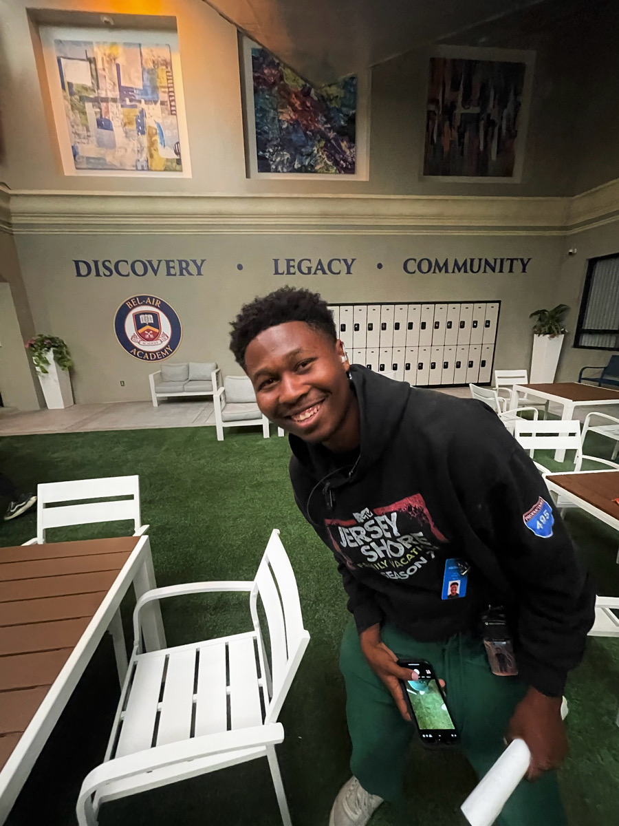 Young man smiling, holding a phone, standing in an indoor space with white chairs and a green carpet.