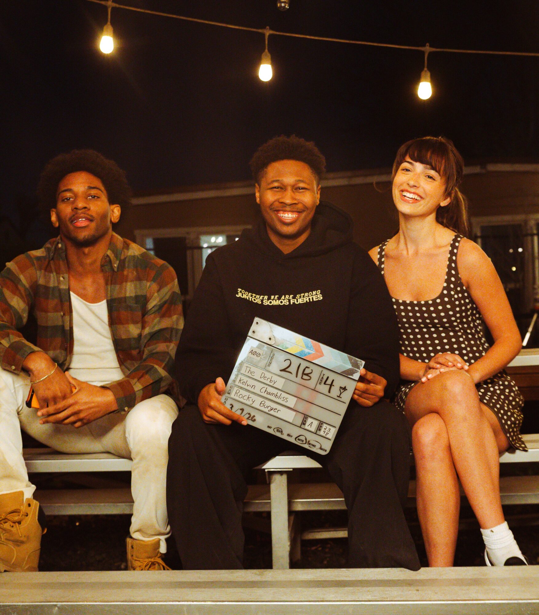 Three people sitting on a bench outdoors at night, smiling, with string lights overhead, one holding a clapperboard.