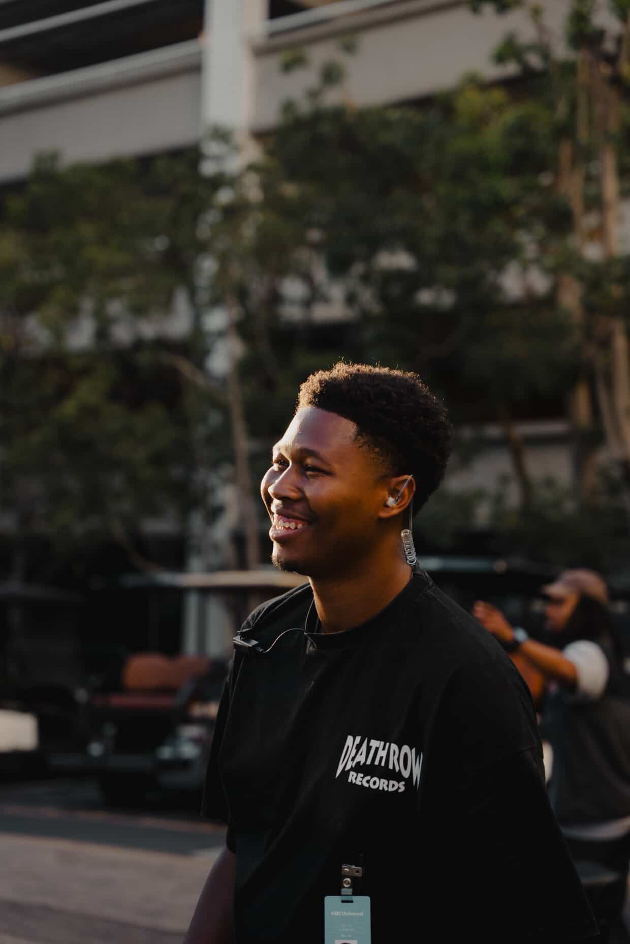 Young man smiling outdoors, wearing a black jacket with a logo, trees and a building in background.