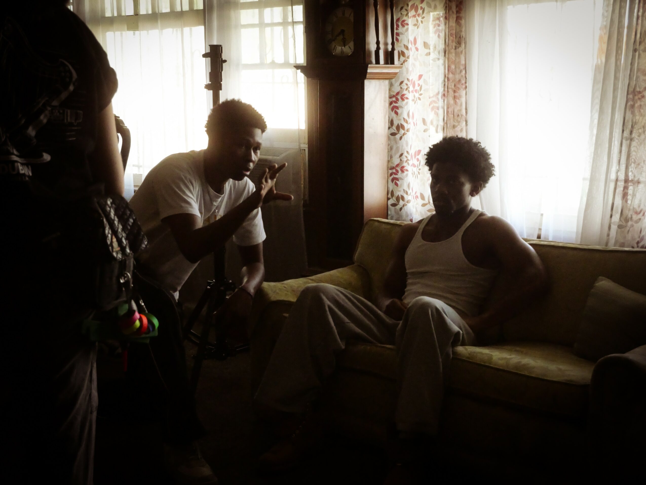 Two people in a dimly lit room, one sitting on a sofa and the other standing, with sunlight coming through curtains.