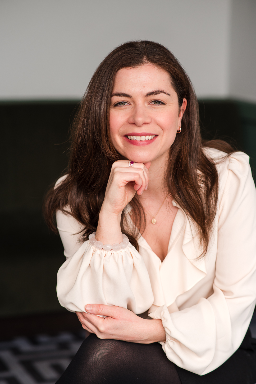 Smiling woman with long brown hair, wearing a white blouse, resting her chin on her hand, in an indoor setting.