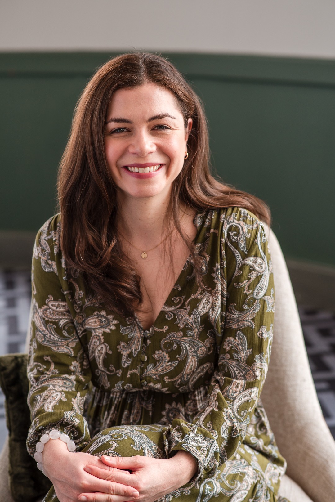 Young woman with long brown hair smiling, wearing a green patterned dress, sitting indoors against a green wall.