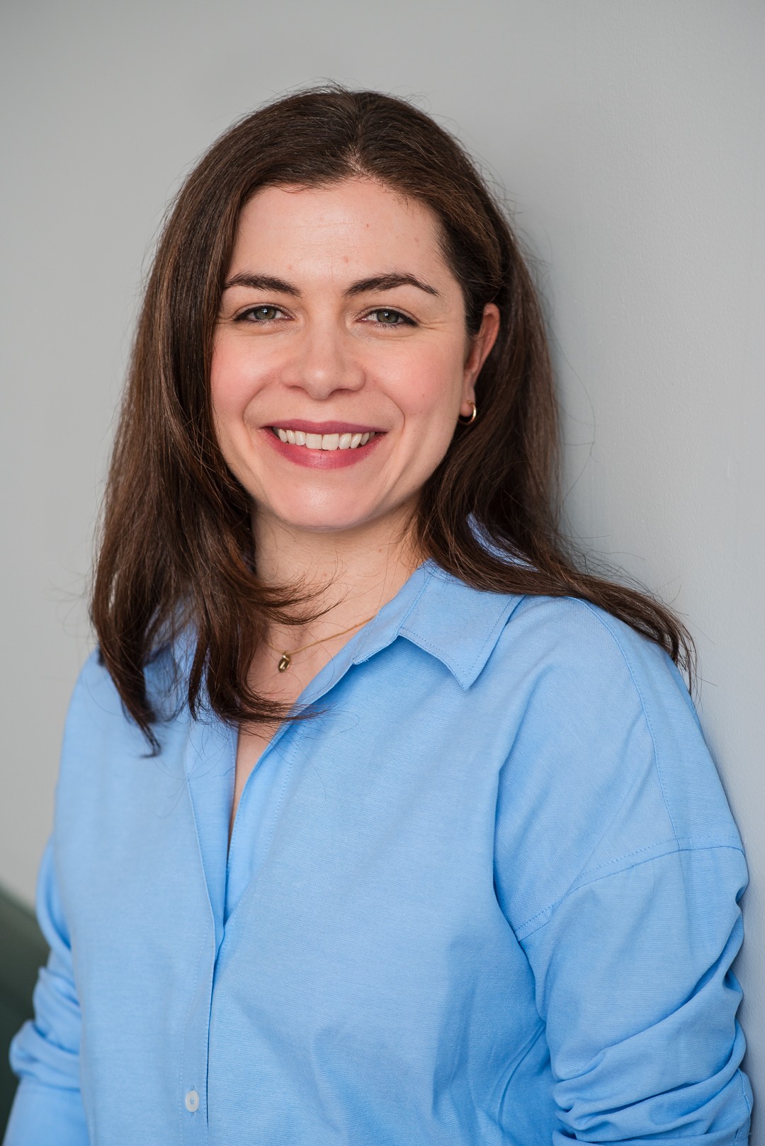 Smiling woman with long brown hair wearing a light blue shirt, standing against a plain background.