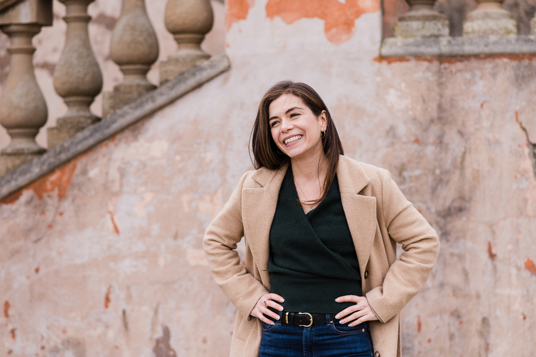 Smiling woman with dark hair in a beige coat standing outdoors against a weathered wall.