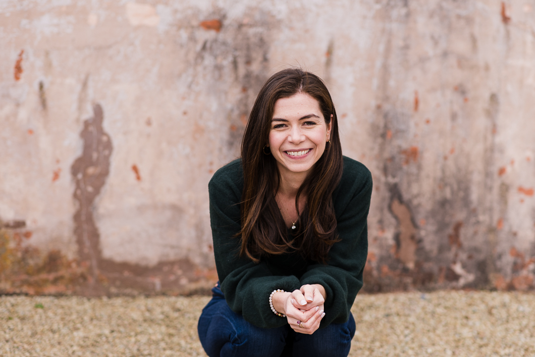 Young woman with long dark hair smiling, standing outdoors against a textured wall, wearing a dark sweater.