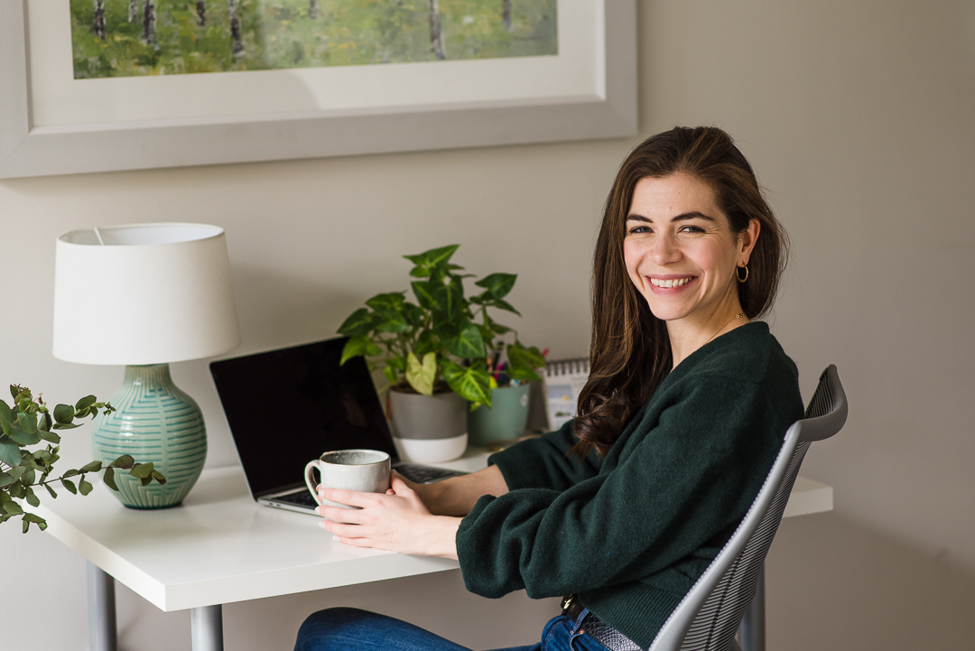 Smiling woman sitting at a desk with a laptop, plants, and a lamp in a bright room.