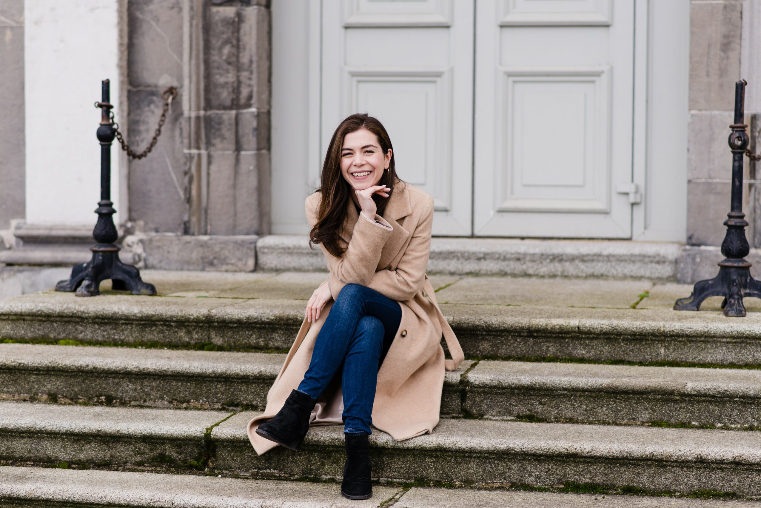 Young woman sitting on steps outside a building, smiling, wearing a beige coat and blue jeans.