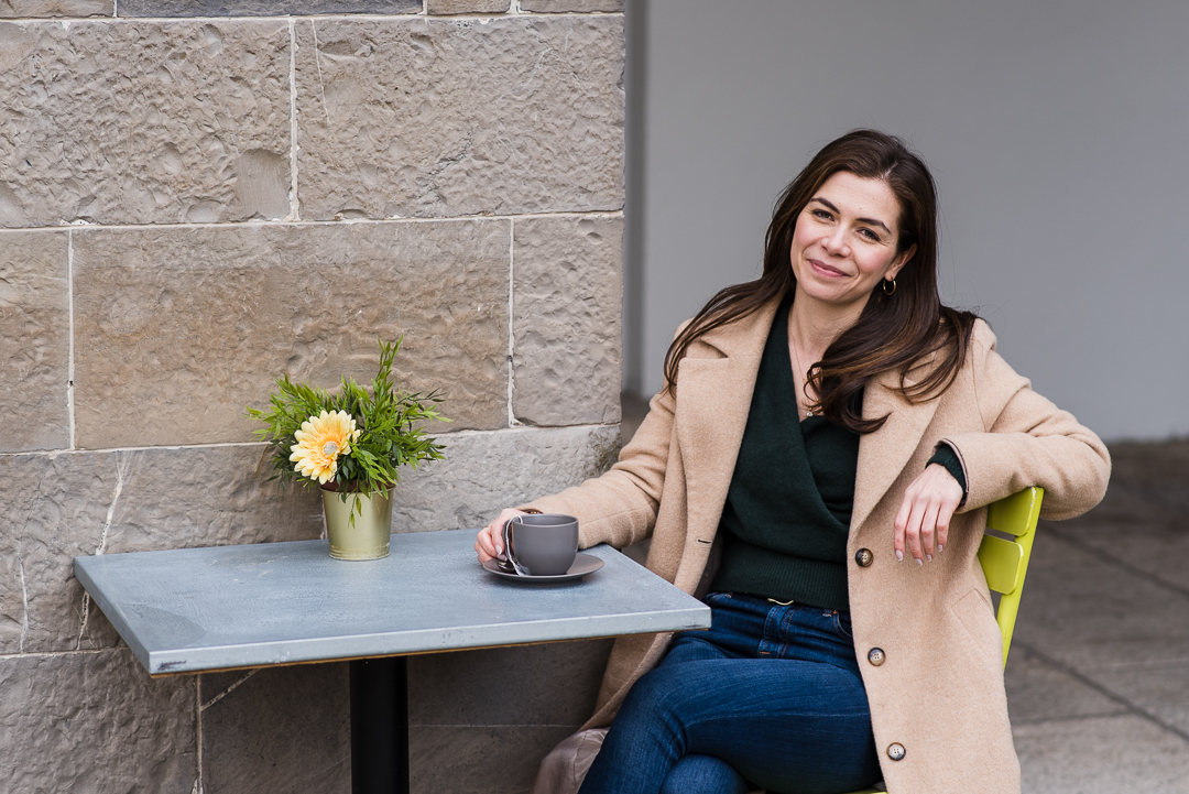 Woman sitting at a table with a potted plant and a cup, smiling, against a stone wall and gray background.