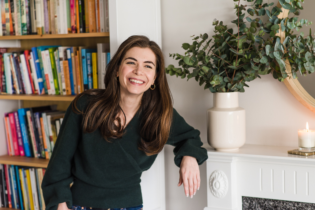 Young woman with long brown hair smiling, sitting in a room with a bookshelf and a plant, wearing a dark green sweater.