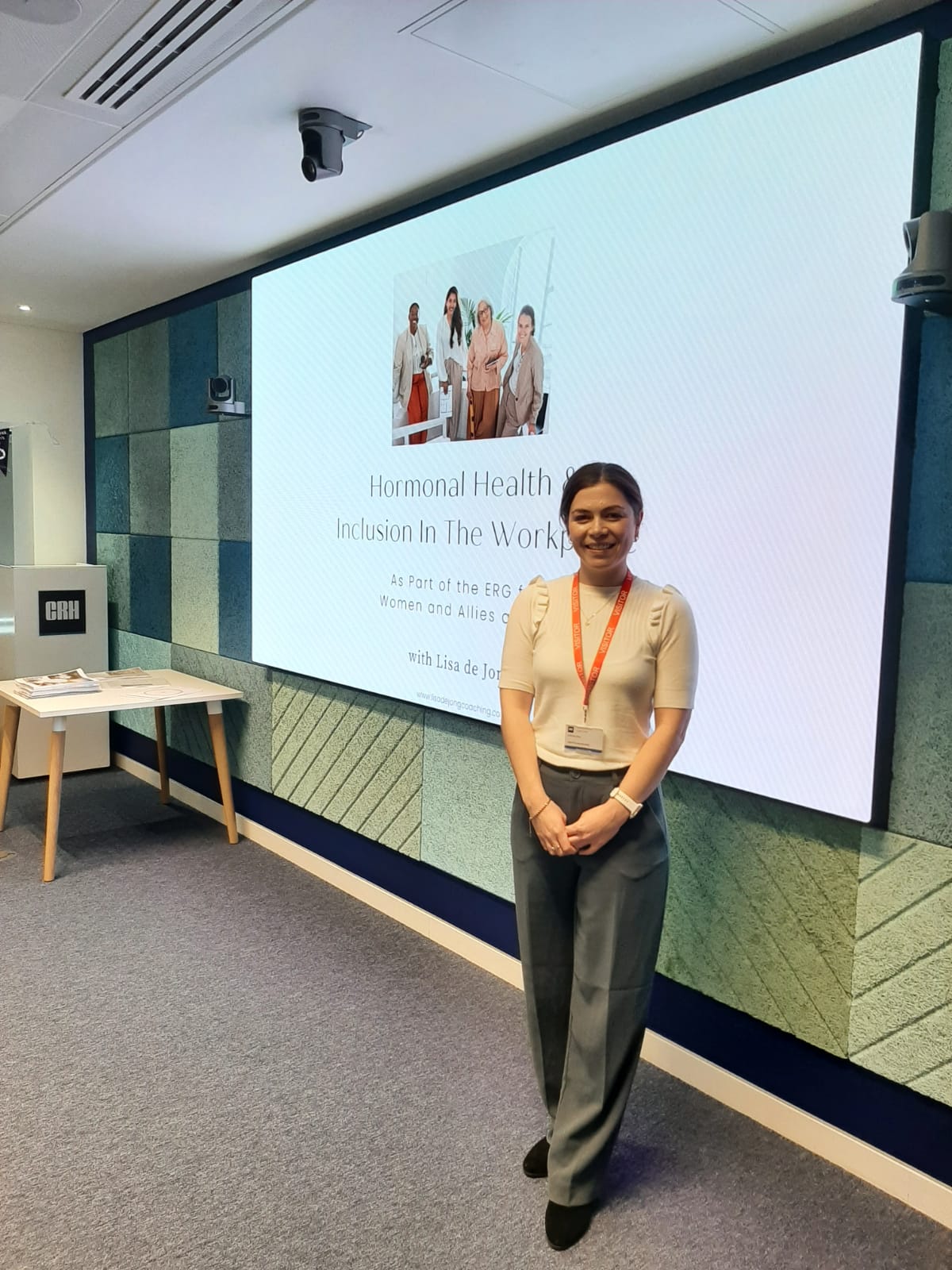Woman standing in front of a presentation screen in a conference room, smiling, wearing a beige top and gray pants.