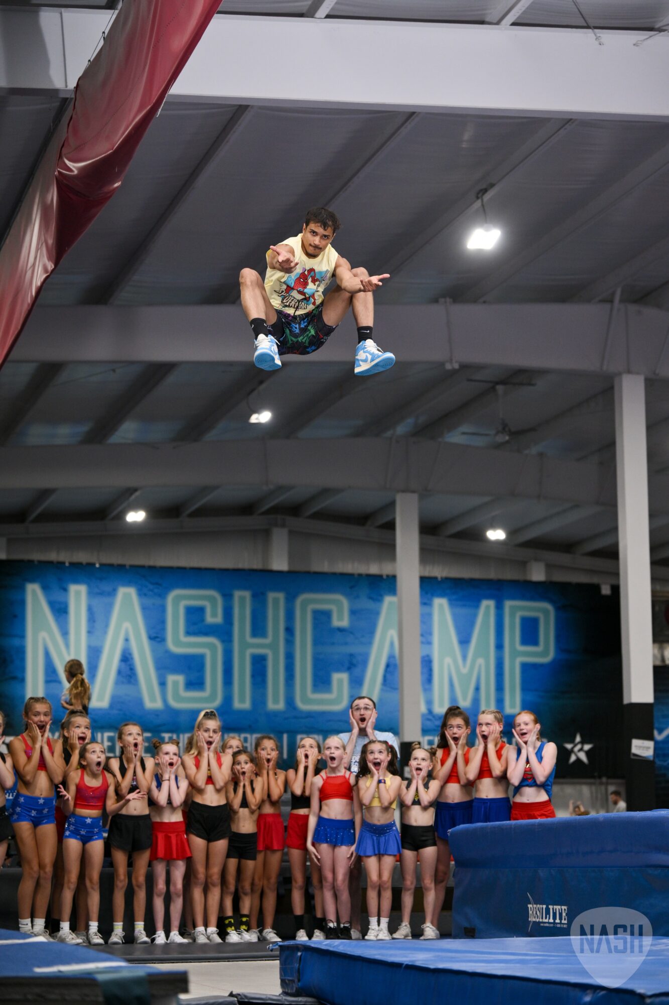 A person is mid-air above a group of cheerleaders at Nash Camp, with a large blue sign in the background.
