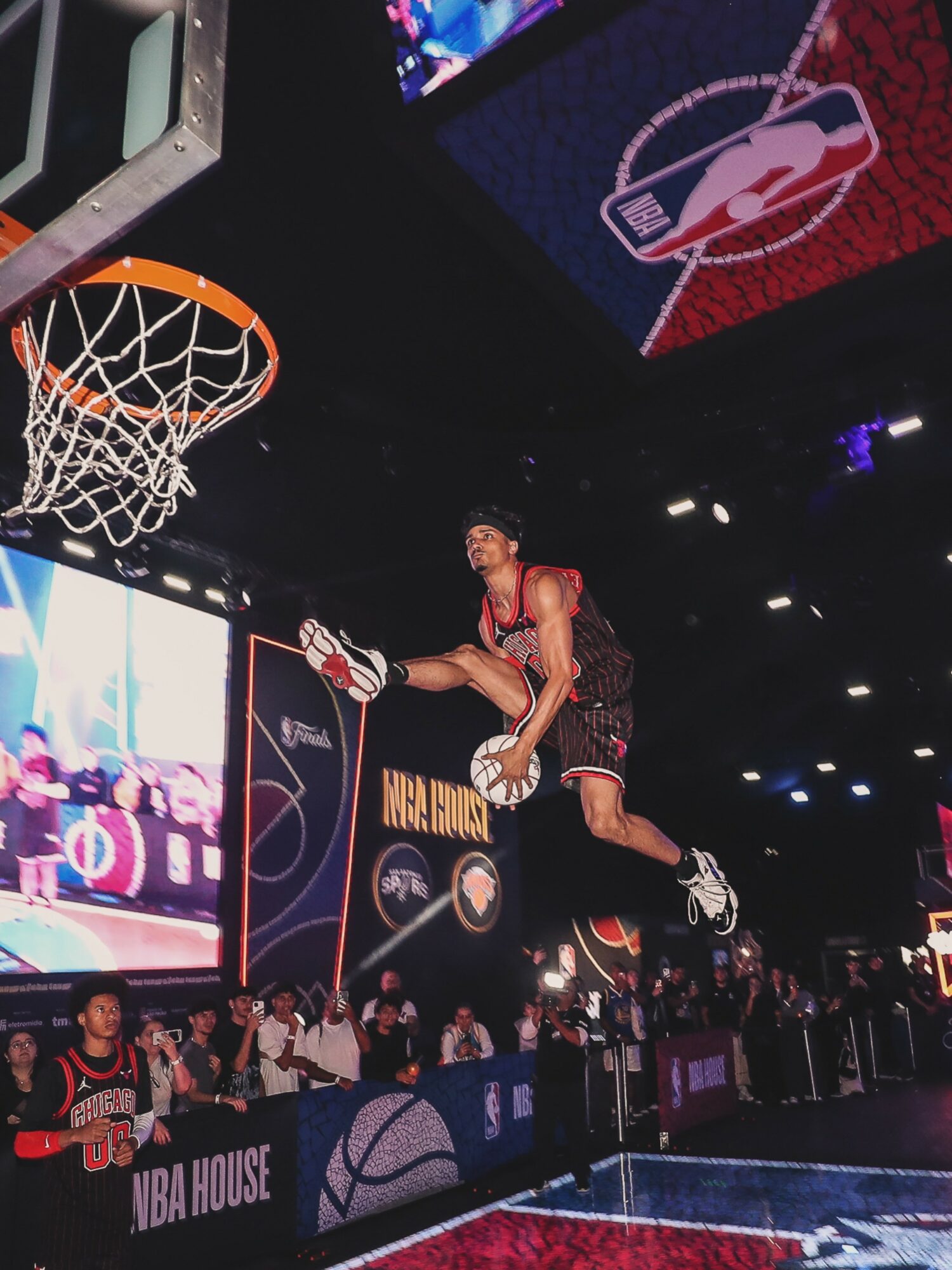 Basketball player mid-air during dunk contest, audience watching, NBA logo on screen, bright arena lights, crowd in background.