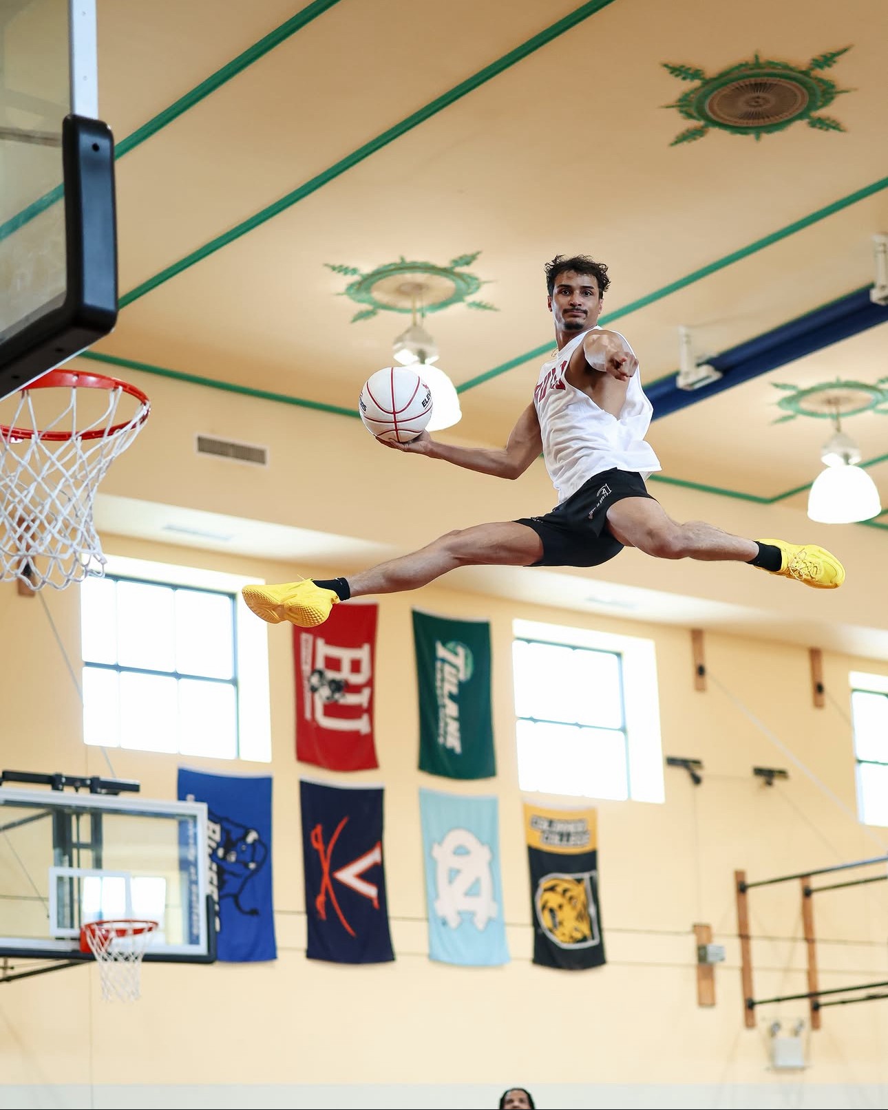 Person performing a split leap in a gymnasium, holding a basketball, with banners hanging on the wall behind.