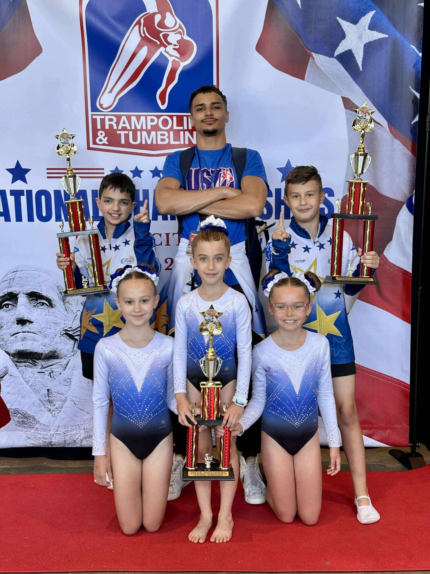 Group of five young gymnasts with medals and trophies, standing with coach in front of a patriotic backdrop.