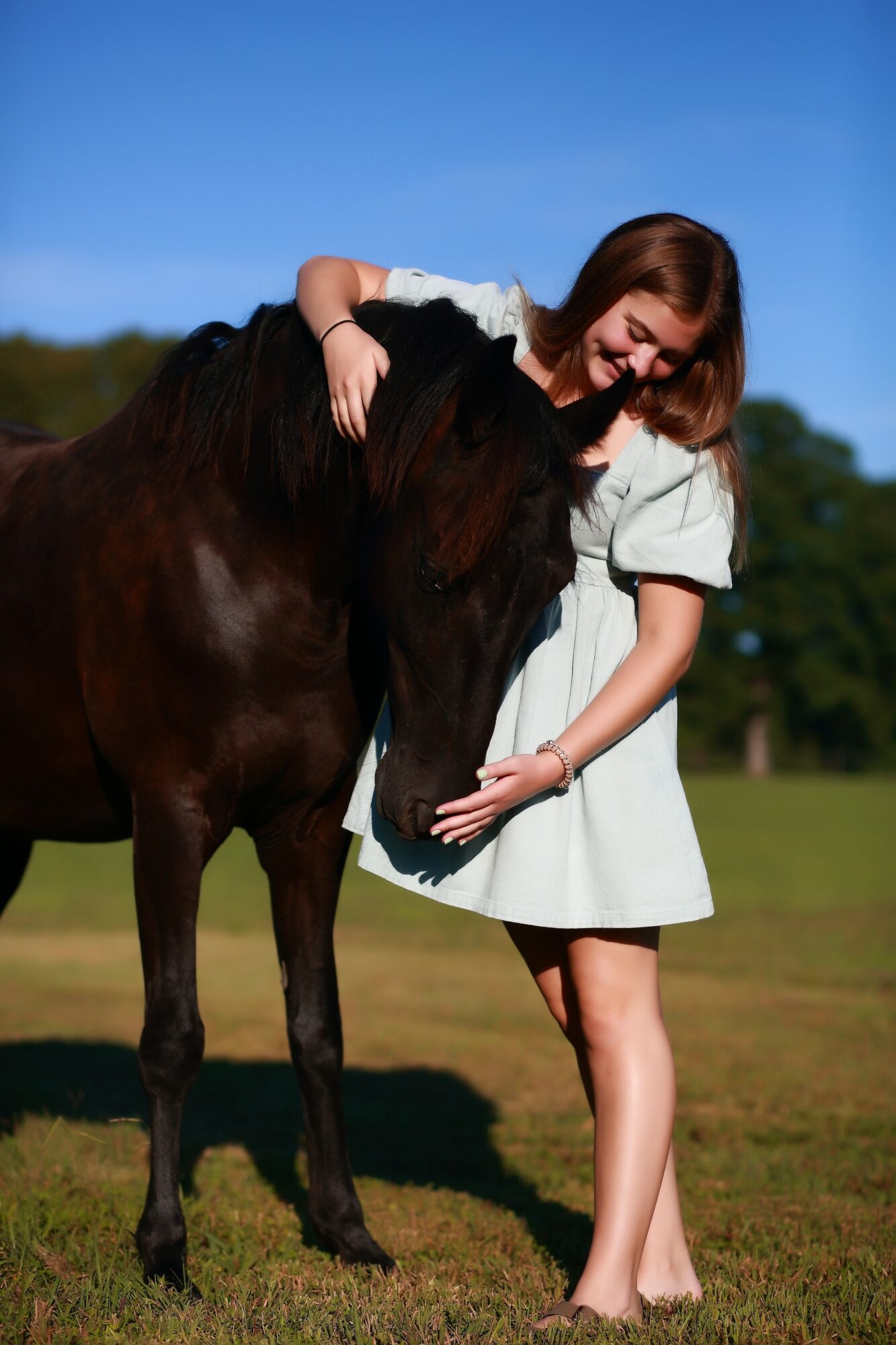 Young woman in a white dress hugging a dark horse outdoors on grass, with trees and blue sky in background.