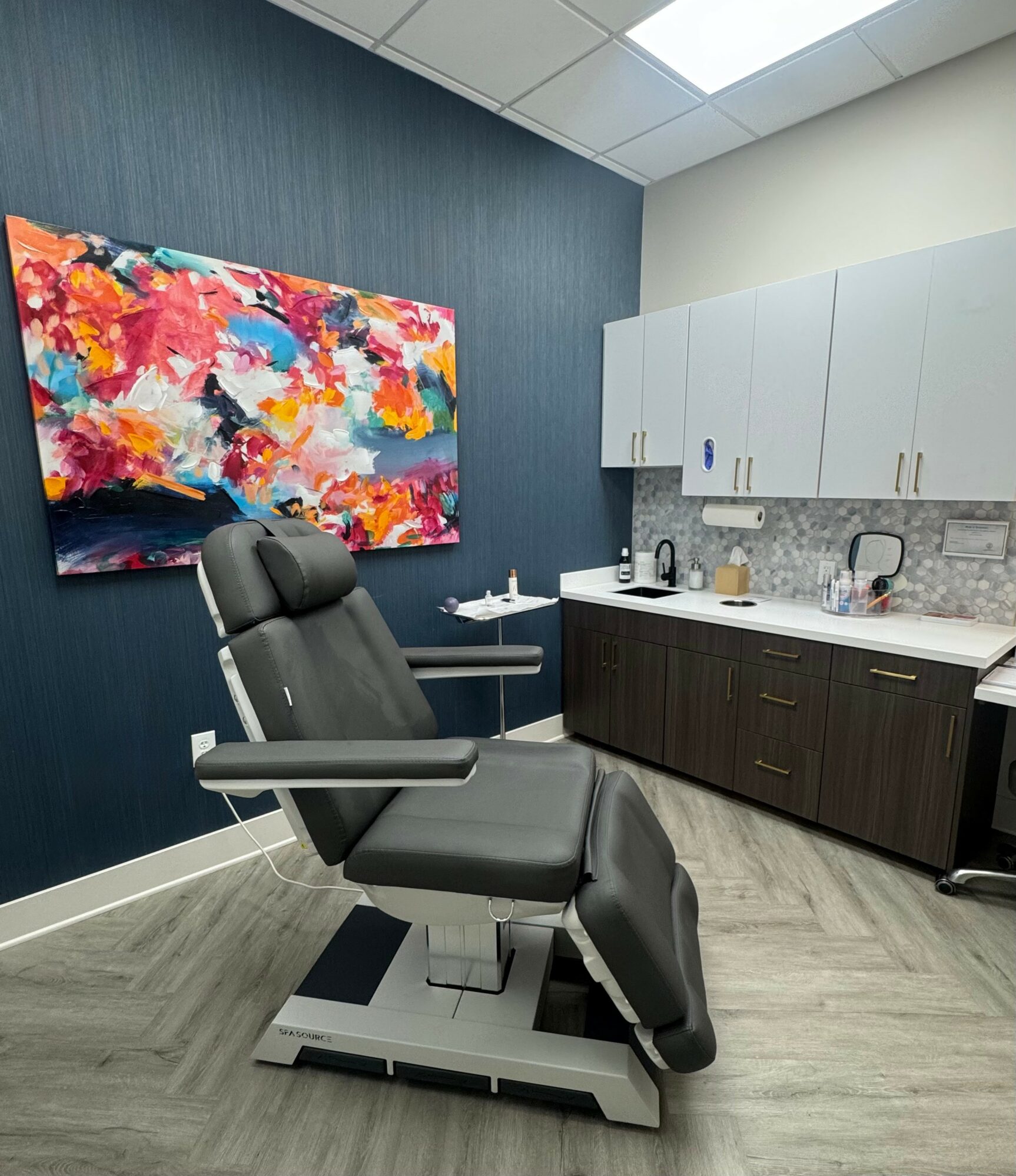Medical examination chair in a room with colorful abstract painting, white cabinets, and a dark wood cabinet, with a gray floor.