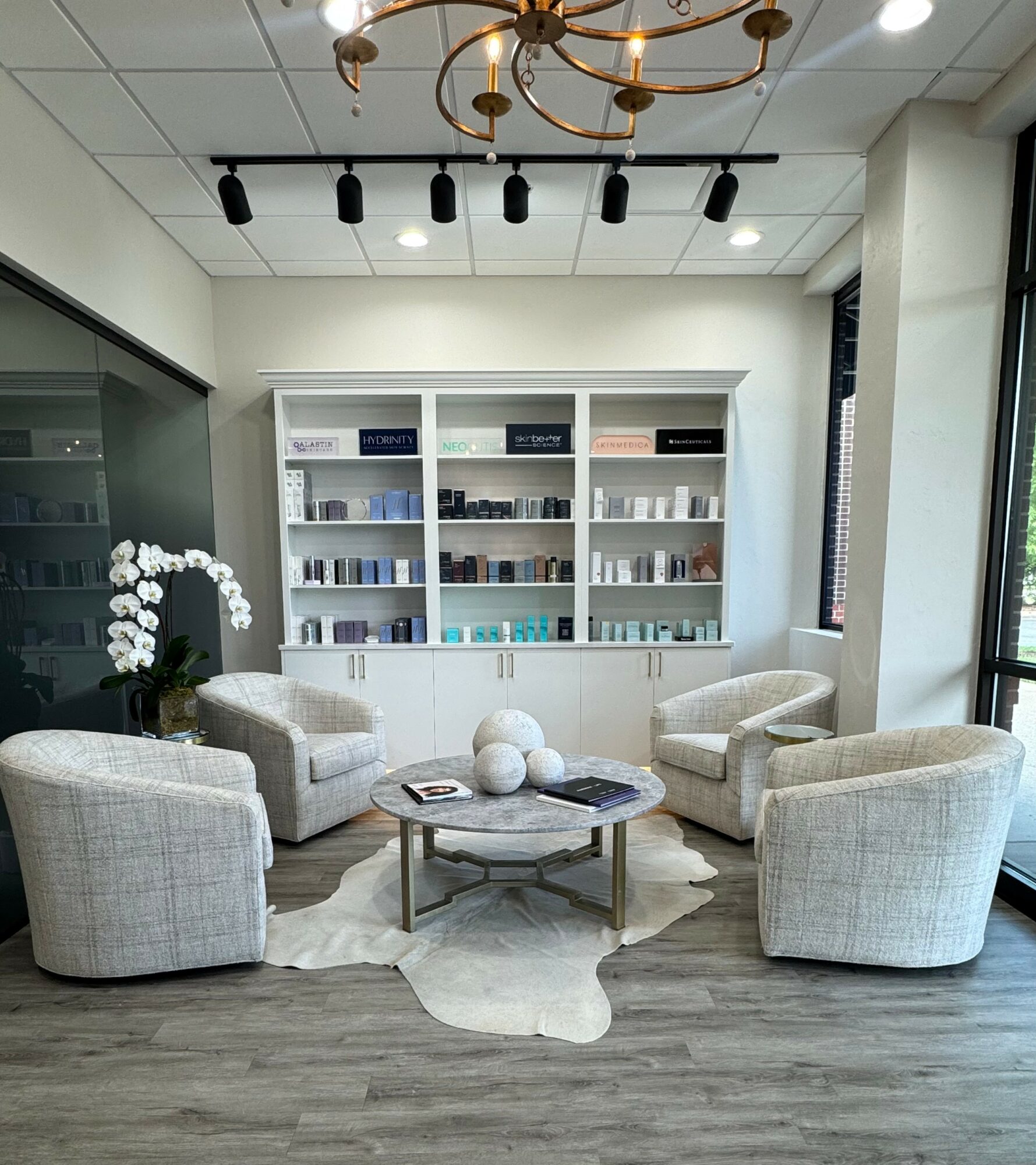 Modern lounge with four armchairs around a round coffee table, bookshelf, and large window, in a bright room.
