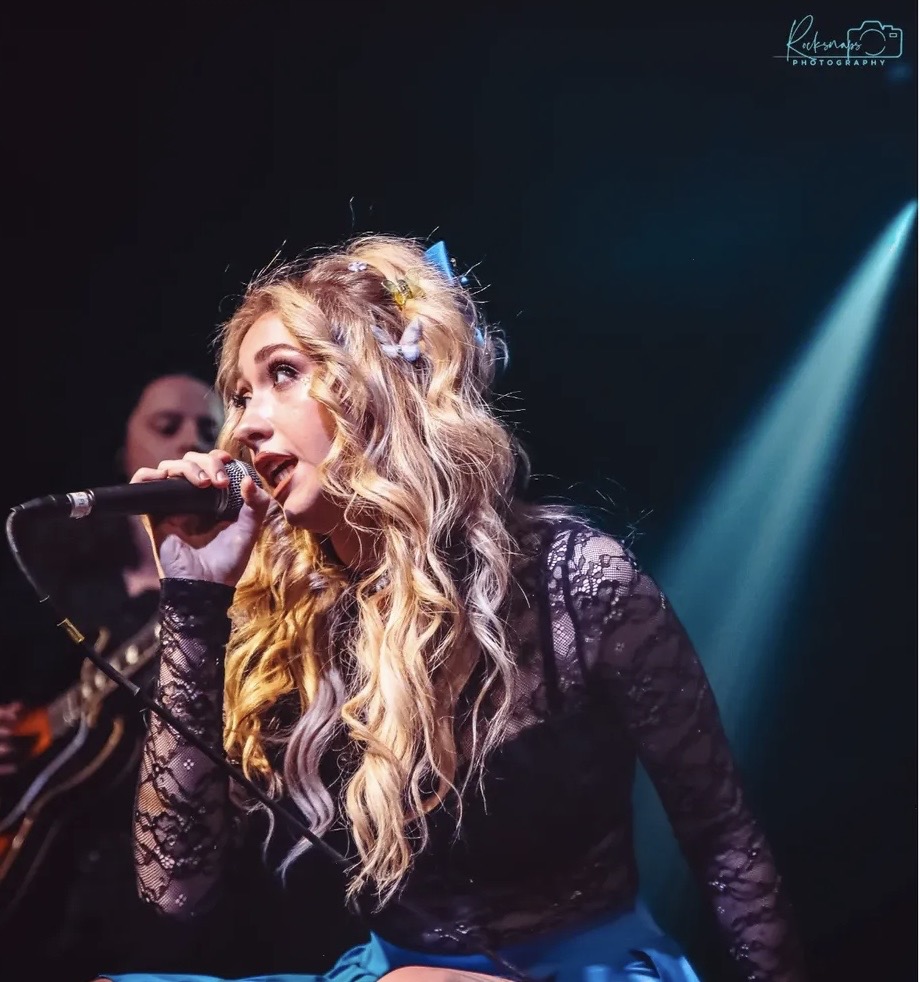 A woman with long, wavy blonde hair sings into a microphone on stage, wearing a black lace top, with stage lighting in background.