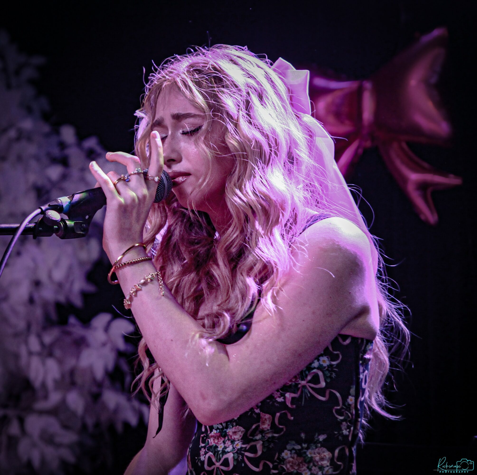 A woman with long, wavy blonde hair sings into a microphone with her eyes closed, wearing a floral dress and jewelry.
