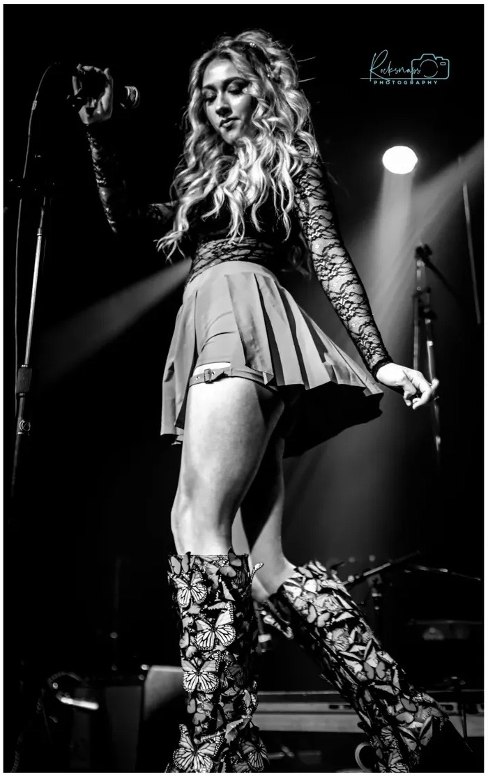 A woman with long curly hair performs on stage, wearing a skirt, lace top, and patterned boots, with stage lights behind her.