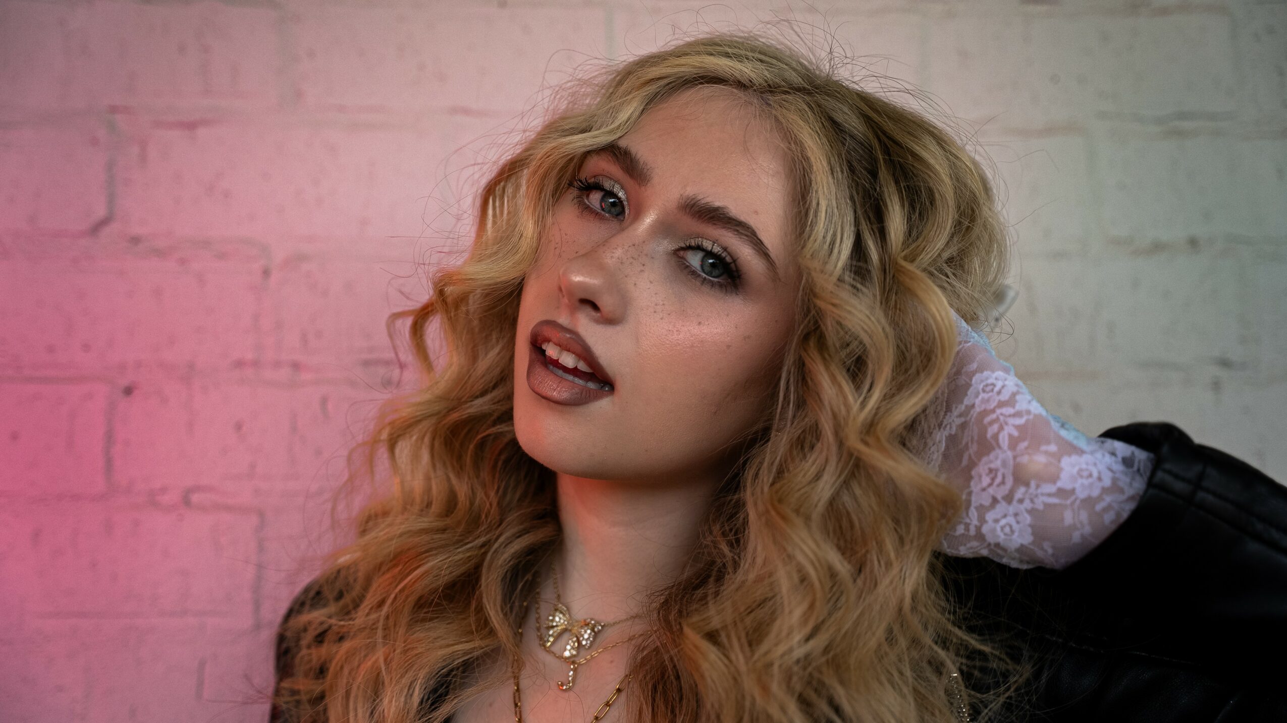 Young woman with wavy blonde hair, makeup, and jewelry, posing against a brick wall with pink lighting.